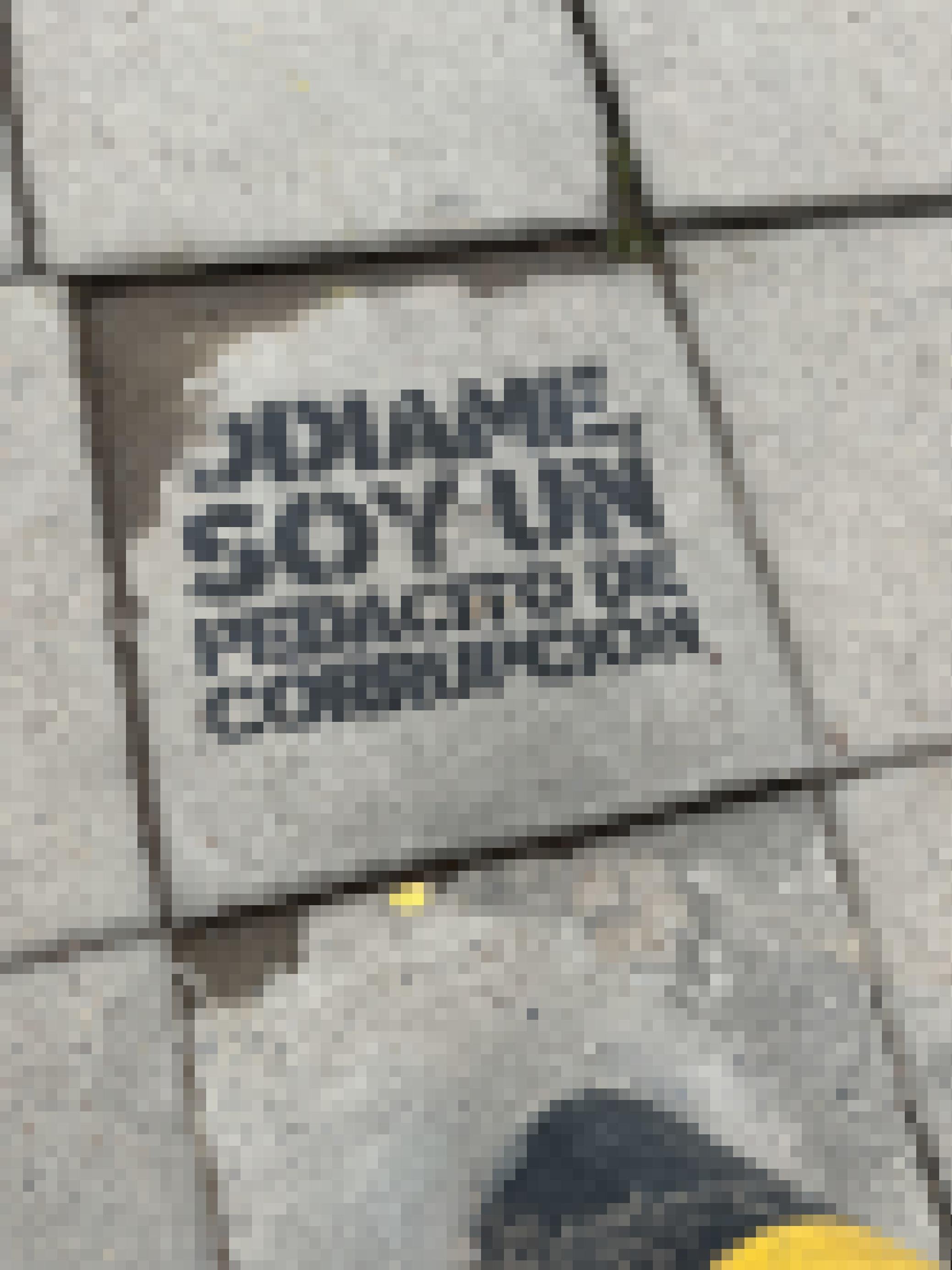 Auf die graue schlichte Bodenplatte eines Gehwegs ist auf Spanisch dieser Text mit einer Schablone gesprüht. Am Rand ist ein Stück Metallpfosten zu erkennen.