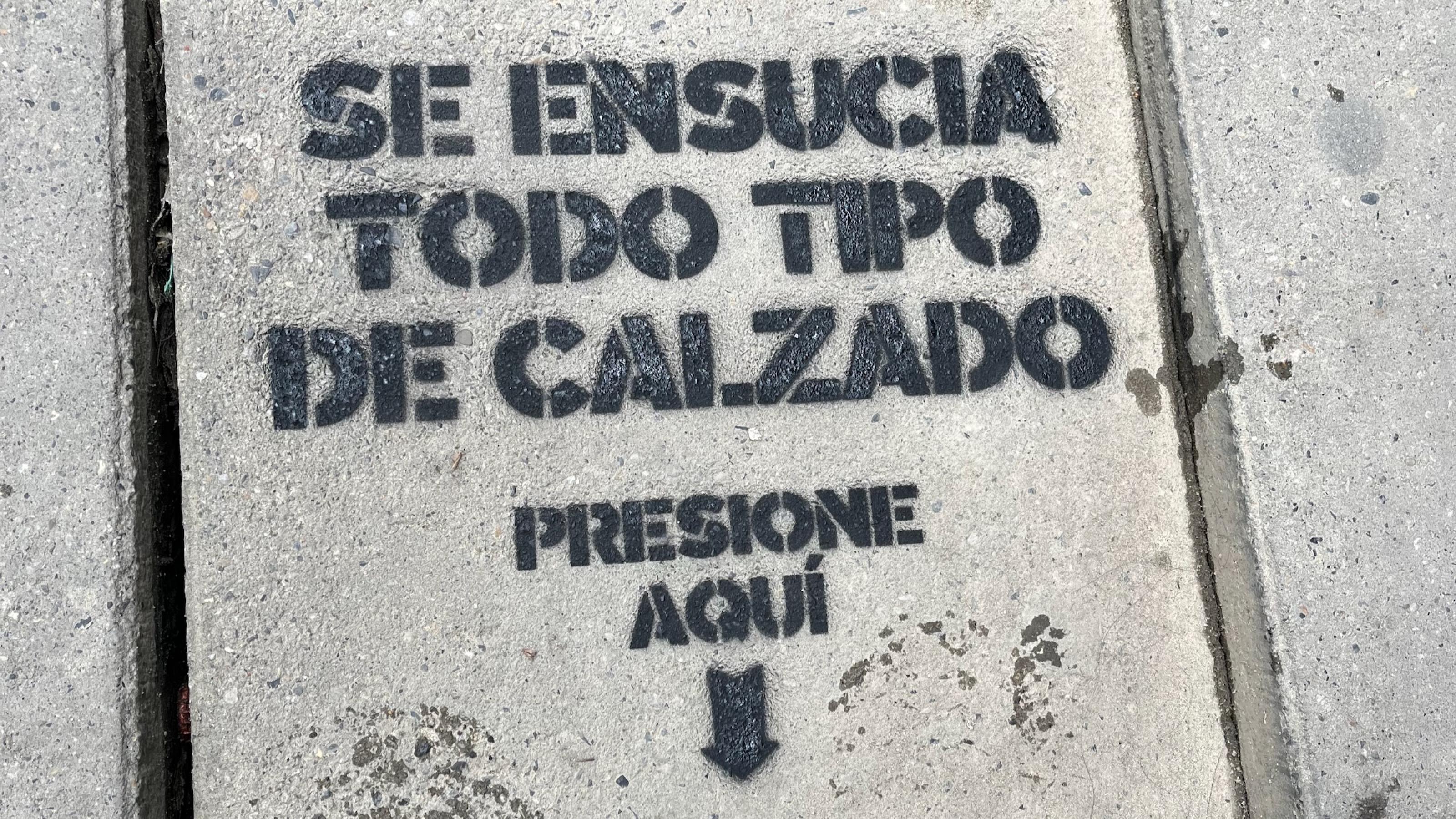 Auf die graue schlichte Bodenplatte eines Gehwegs ist auf Spanisch dieser Text mit einer Schablone gesprüht. Am Ende zeigt ein Pfeil auf die Stelle, auf die man treten soll (was auch an zwei feuchten echten Schuhabdrücken zu erkennen ist).