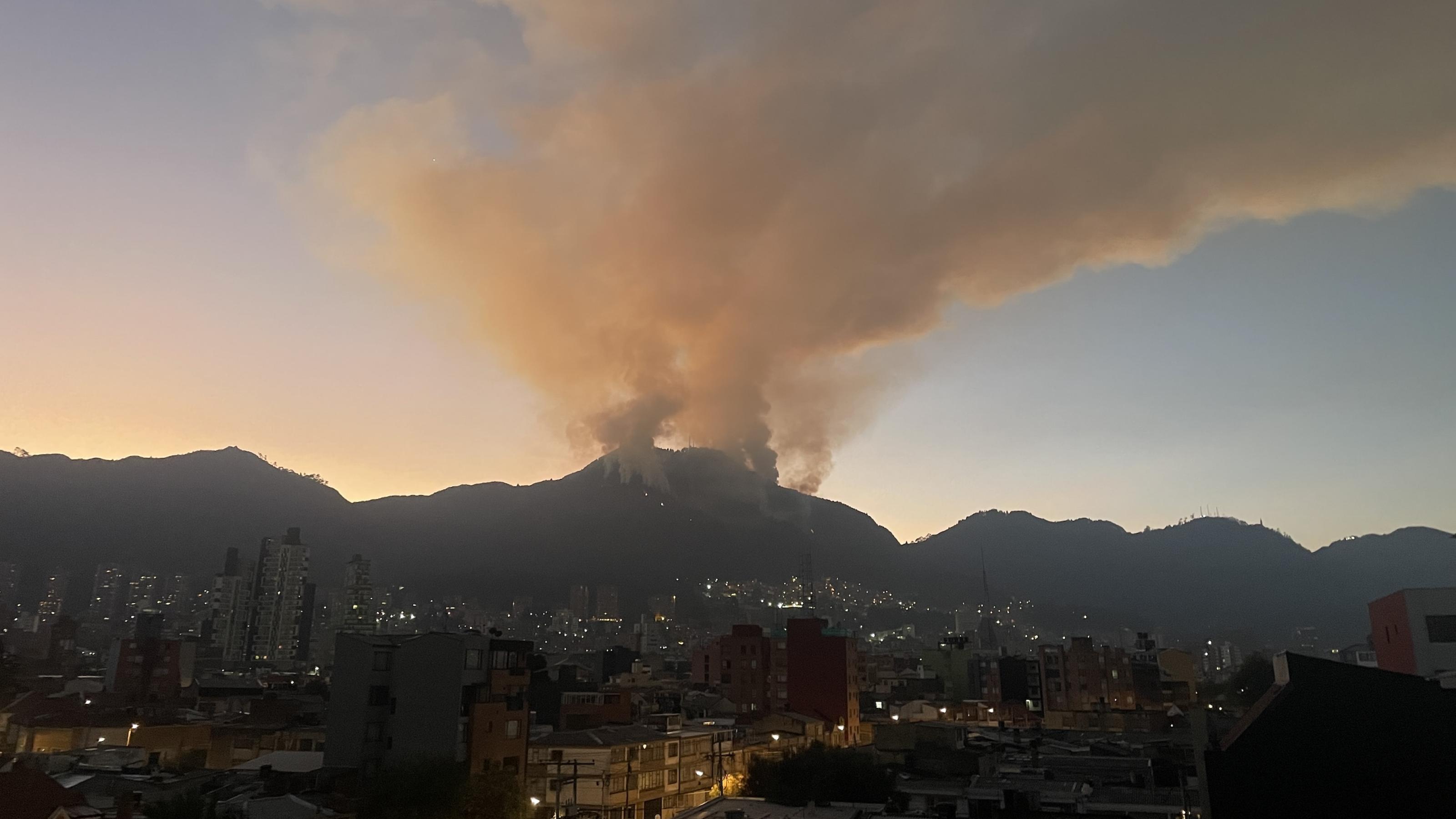 Stadtpanorama von Bogotá mit Hochhäusern in der Dämmerung. Im Hintergrund eine Bergkette. Vom Gipfel in der Mitte zieht ein Rauchkegel in die Höhe. Das sieht aus wie bei einem Vulkan.