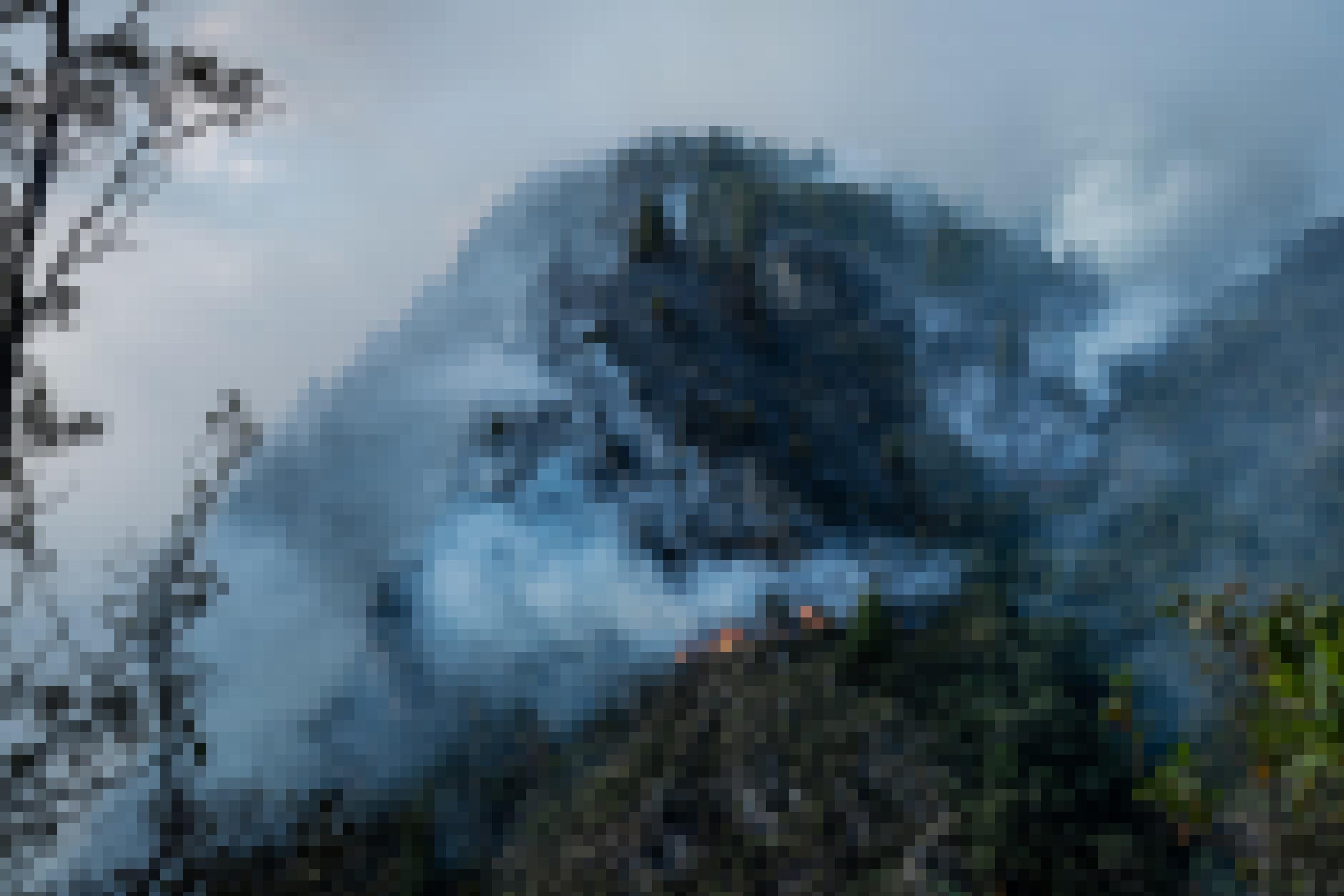 Sicht von oben auf einen mit Nadelbäumen bewachsenen Berg, der steil aus einer Bergkette ragt. Er ist vor lauter Rauch nur teils zu sehen. An mehreren Stellen Flammen.