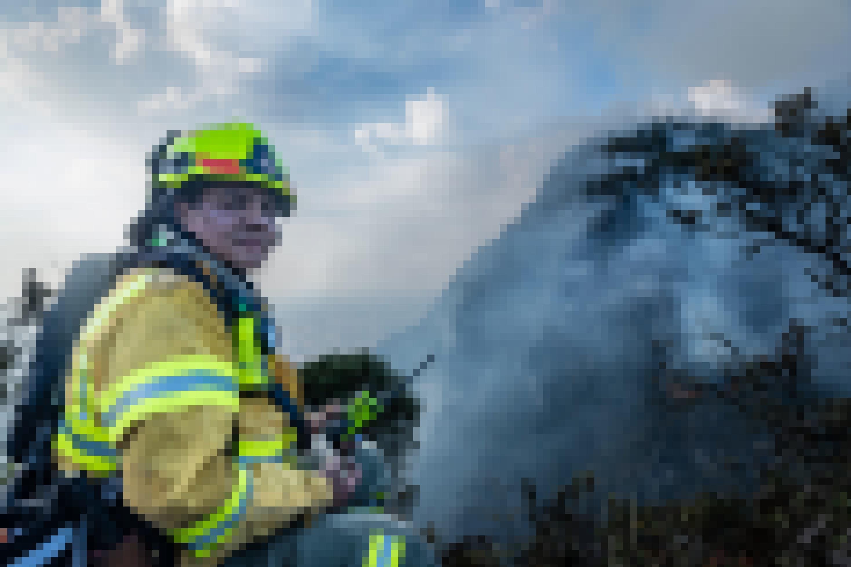 Ein Feuerwehrmann in Schutzkleidung (Helm, Lampe, Funkgerät, Rucksack) blickt mit verrußtem Gesicht und die Kamera. Im Hintergrund ein von Rauchschwaden fast komplett verhüllter Berg.