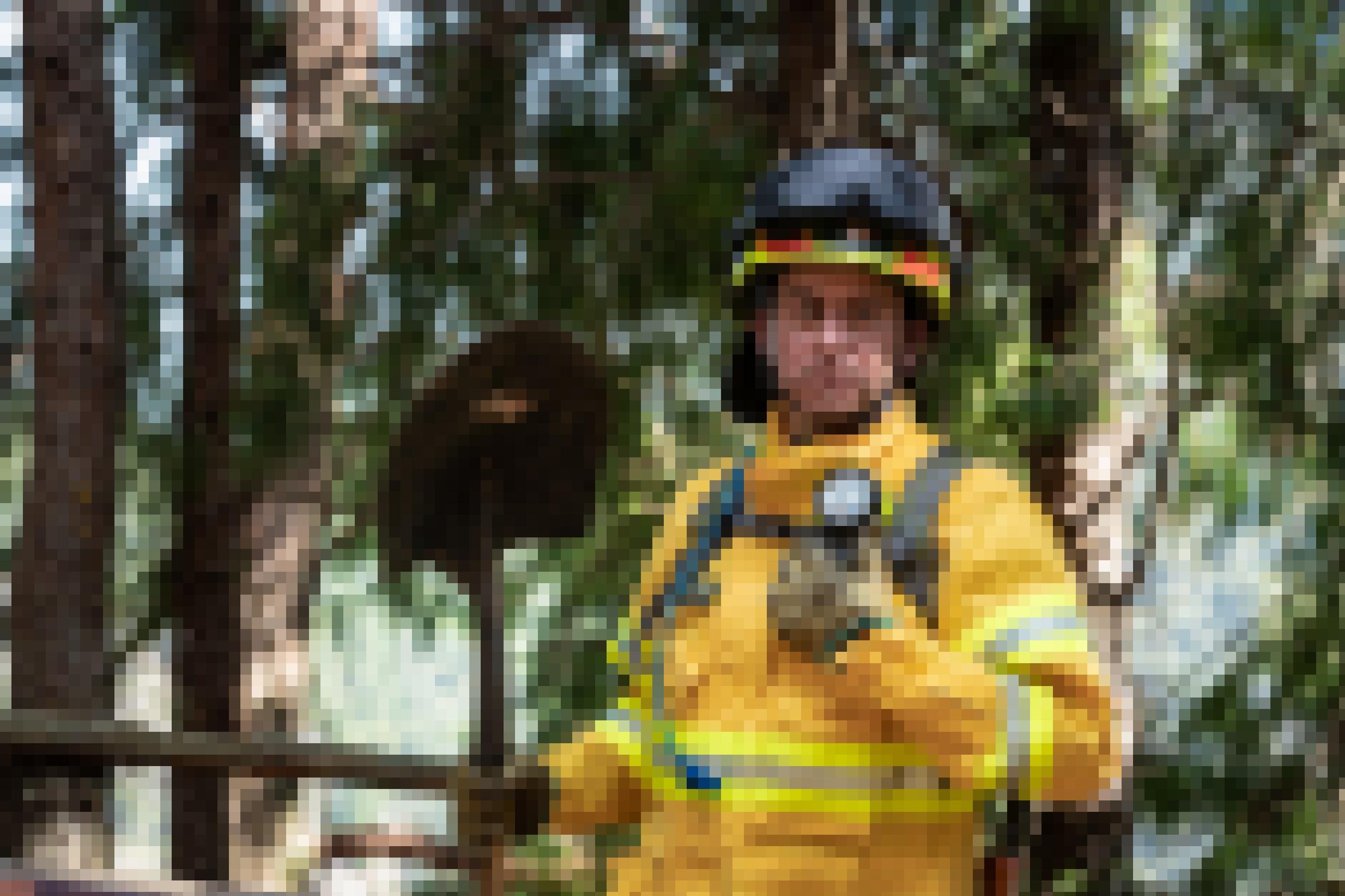 Feuerwehrmann in gelber Einsatzkleidung, Helm, verrußten Handschuhen, Spaten in der Hand im Kiefernwald. Er sieht müde aus.