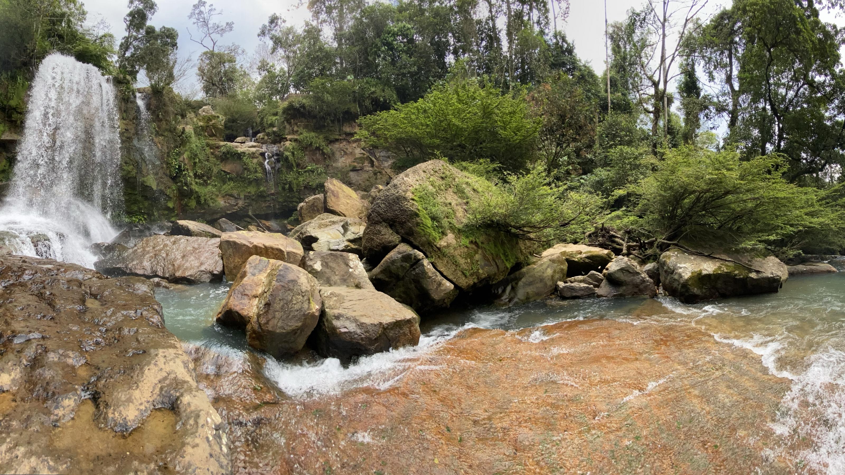 Links ein Wasserfall, der in einen schmalen, klaren Fluss mündet, dahinter Felsen.