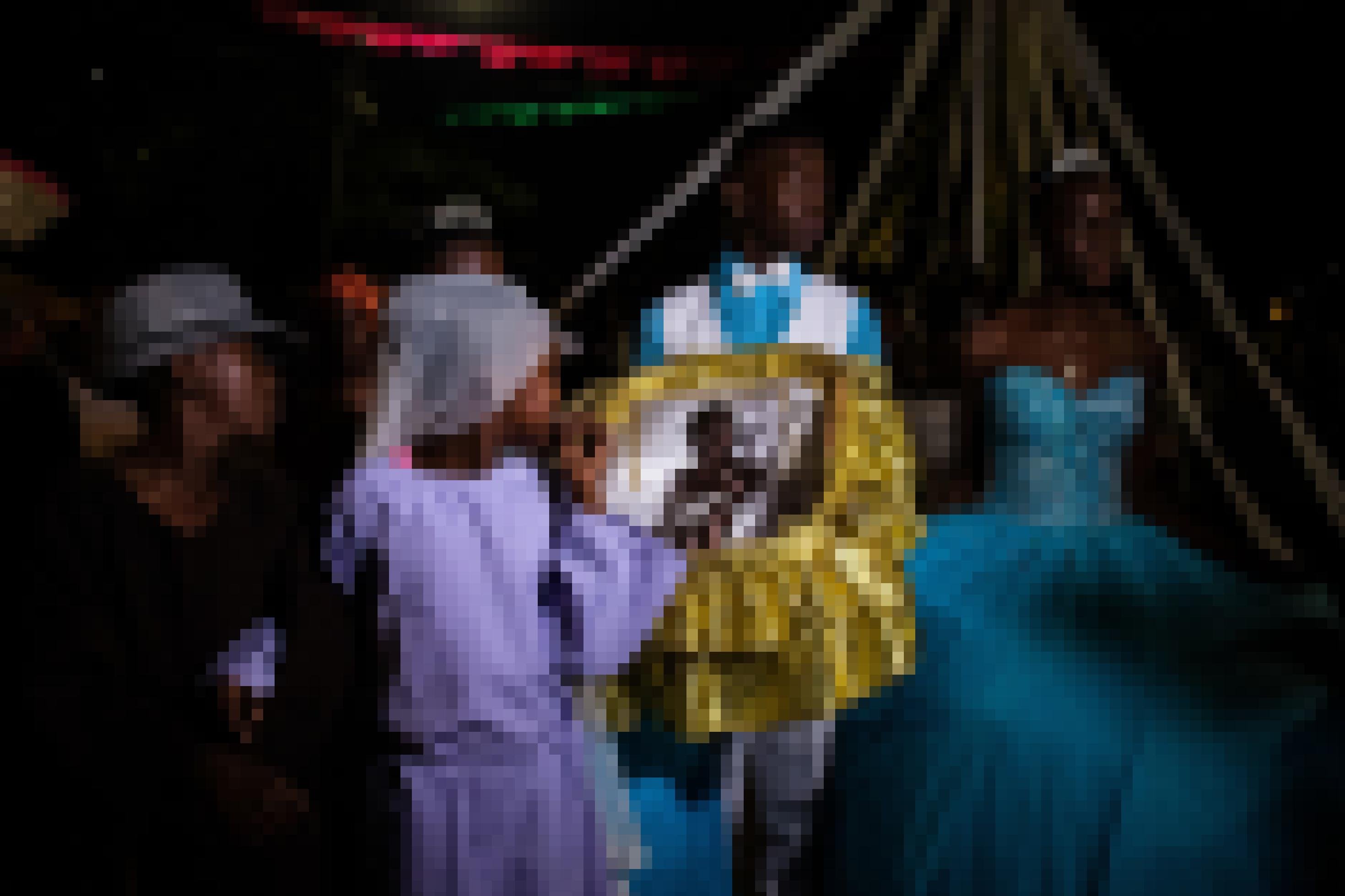 Zwei Schwarze Jugendliche tragen leuchtendes hellblau. Er einen Anzug, sie ein Ballkleid mit Diadem. Halb verdeckt sind drei jüngere Kinder zu sehen: Ein Mädchen trägt Diadem, das andere ein Kopftuch, der Junge einen Hut. In ihrer Mitte tragend die fünf ein mit Goldstoff ausgeschlagenes Körbchen mit einer schwarzen Babypuppe.