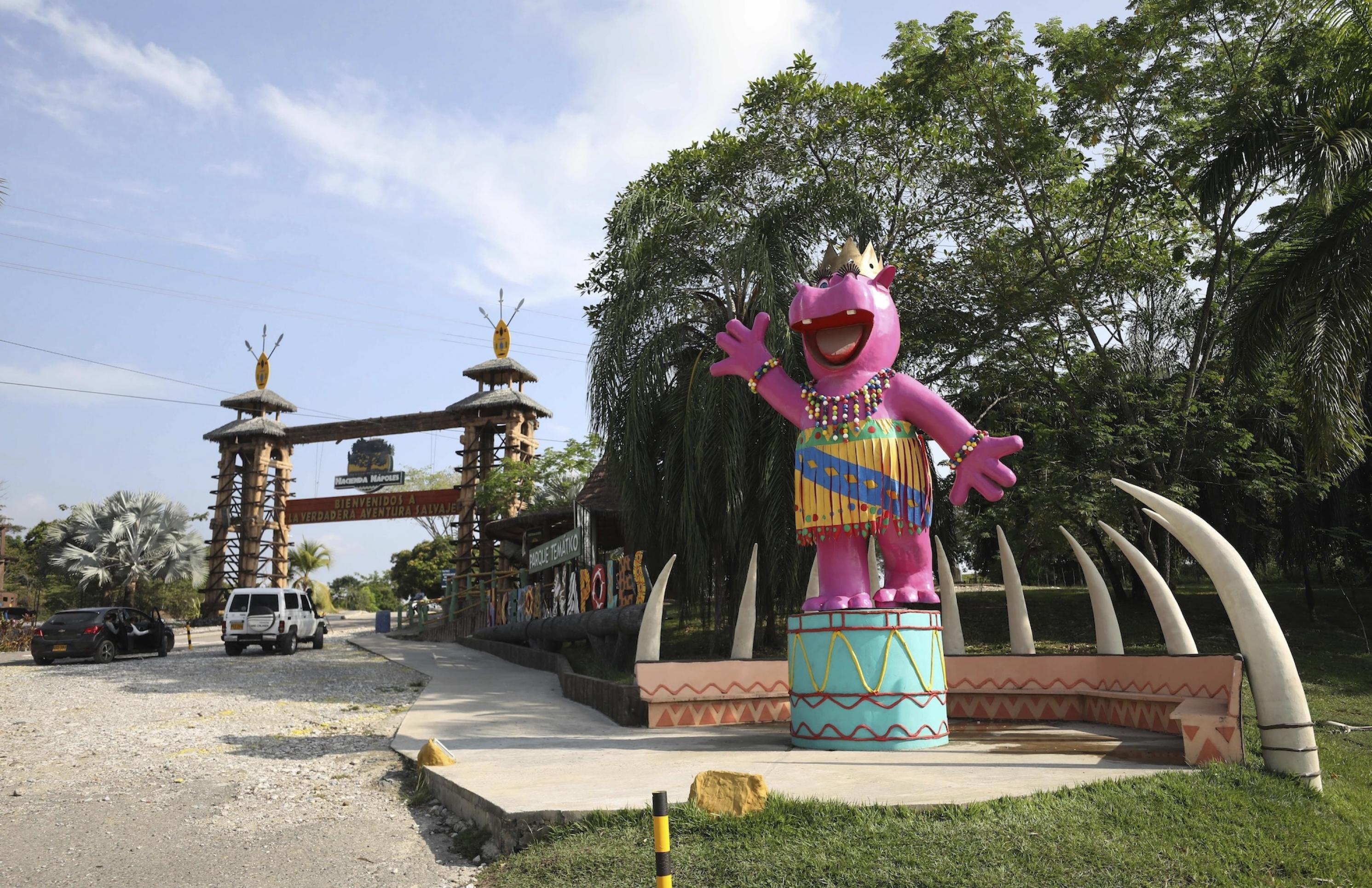 Ein riesiges rosa Beton-Nilpferd mit buntem Röckchen und Armbändern in afrikanischem Stil hebt die rechte Hand zum Gruß und lacht. Es steht auf einem trommelähnlichen, türkisen Podest. Hinter dem Hippo ist wie ein Zaun ein Halbrund, das aus künstlichen Elefanten-Stoßzähnen besteht. Im Hintergrund ist ein Tor zu sehen, das an einen Safari-Park erinnert. Ein Jeep und ein Auto nähern sich dem Tor. Überm Tor und daneben auf der Wiese stehen Schilder mit der Aufschrift „Hacienda Nápoles“.