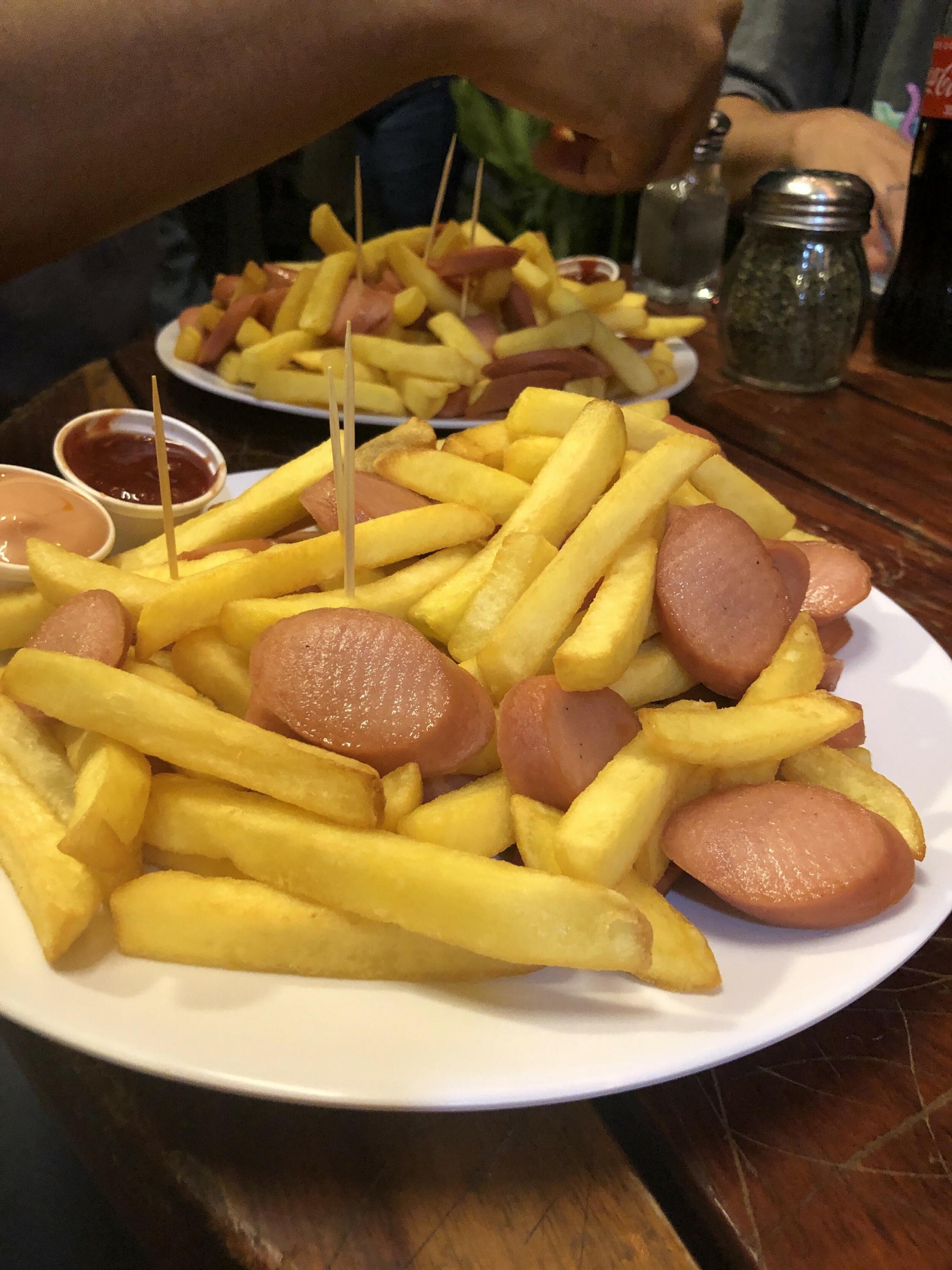 Ein Teller Pommes mit schräg geschnittenen und leicht angebratenen Wurststücken, die wie Wiener aussehen. Darin stecken mehrere Zahnstocher, am Tellerrand sind zwei Plastikbecherchen mit Saucen.