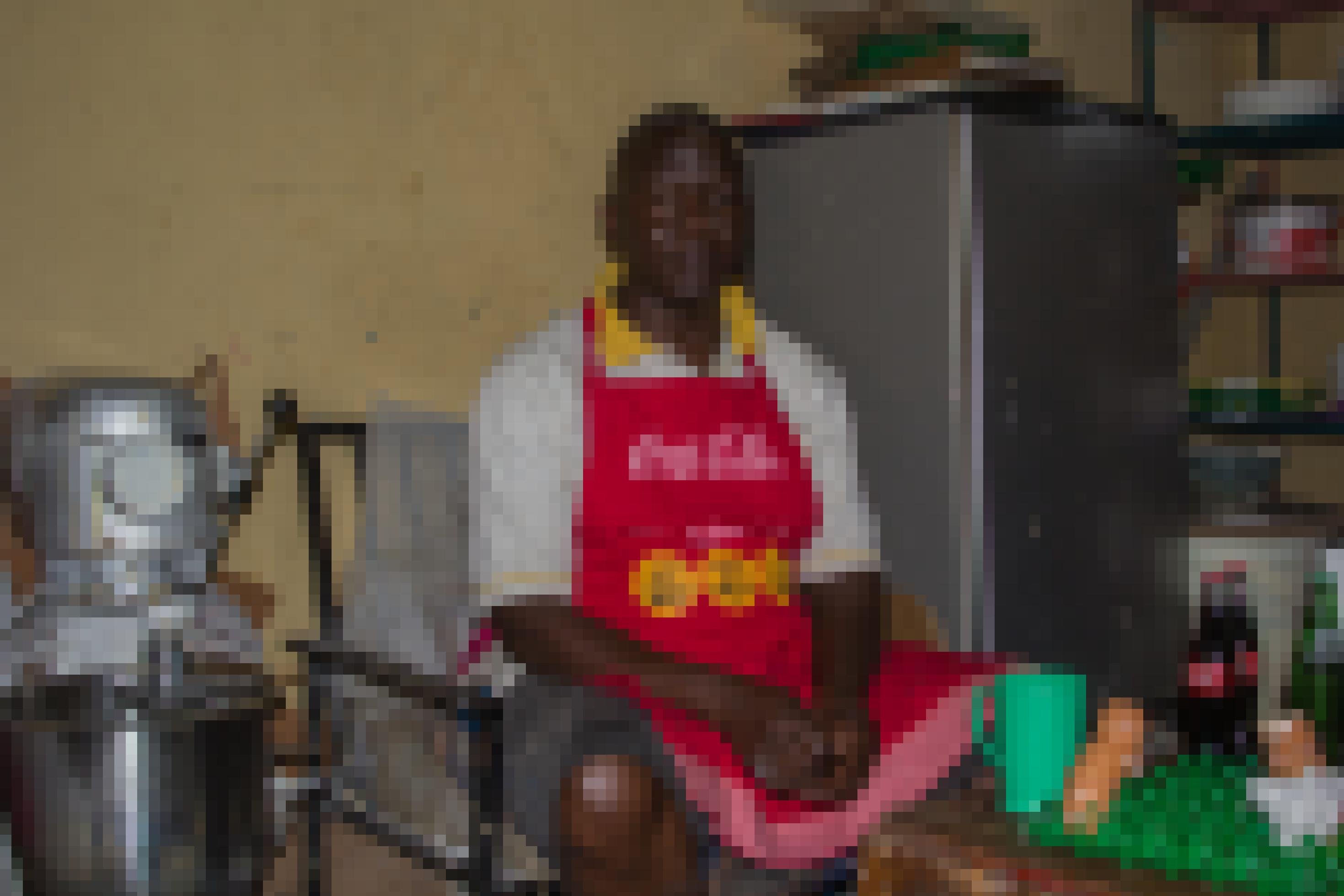 Ein Mann mit roter Schürze sitzt auf einem Stuhl in einem engen Raum. Neben ihm befindet sich ein Kühlschrank und eine Rührmaschine.