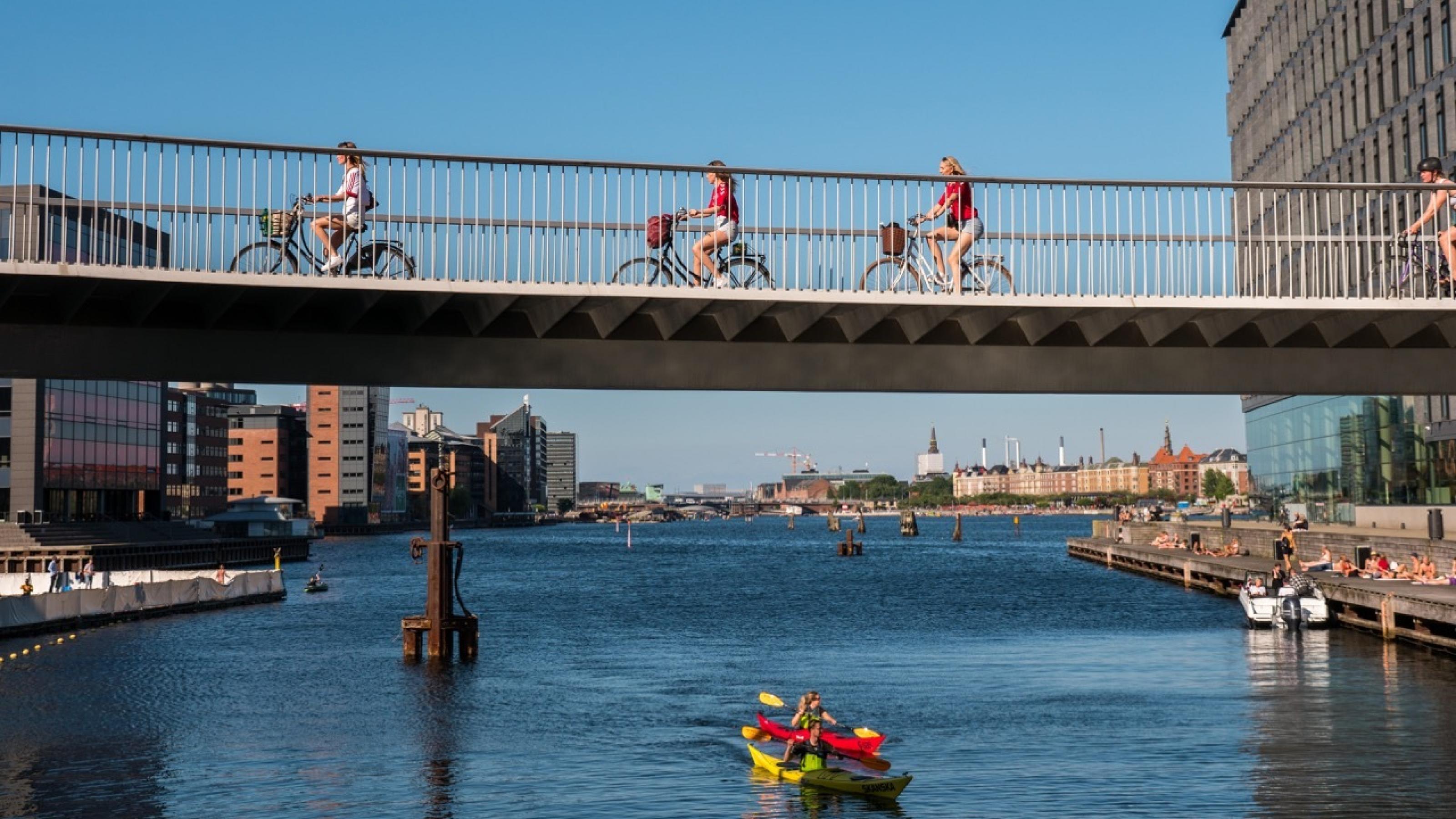 Zu sehen sind Radfahrer auf einer Brücke über ein Gewässer, in sattem Sonnenlicht.