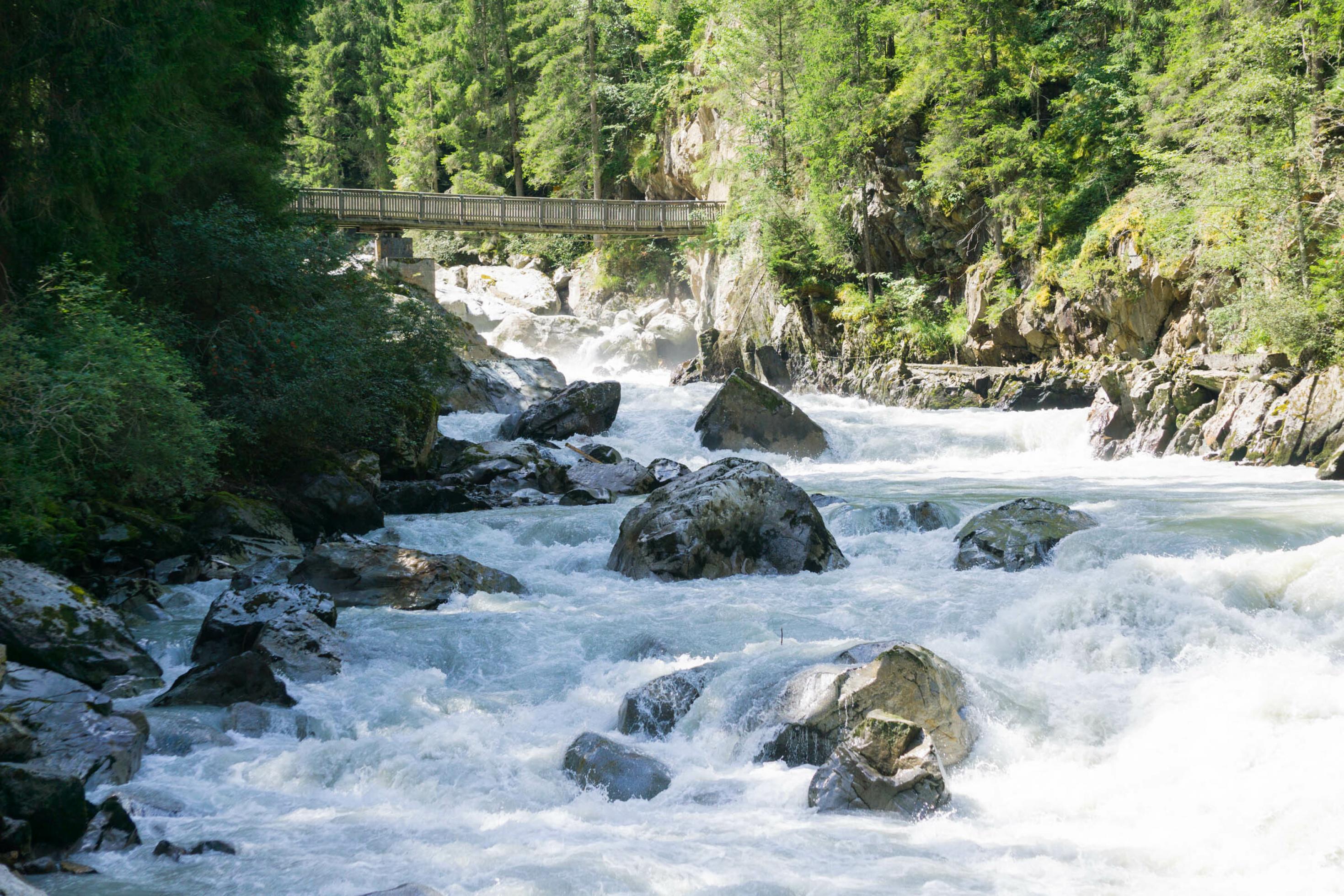 Gletscherbach im Wald mit Felsen und weiß schäumendem Wasser, im Hintergrund eine schmale Holzbrücke.