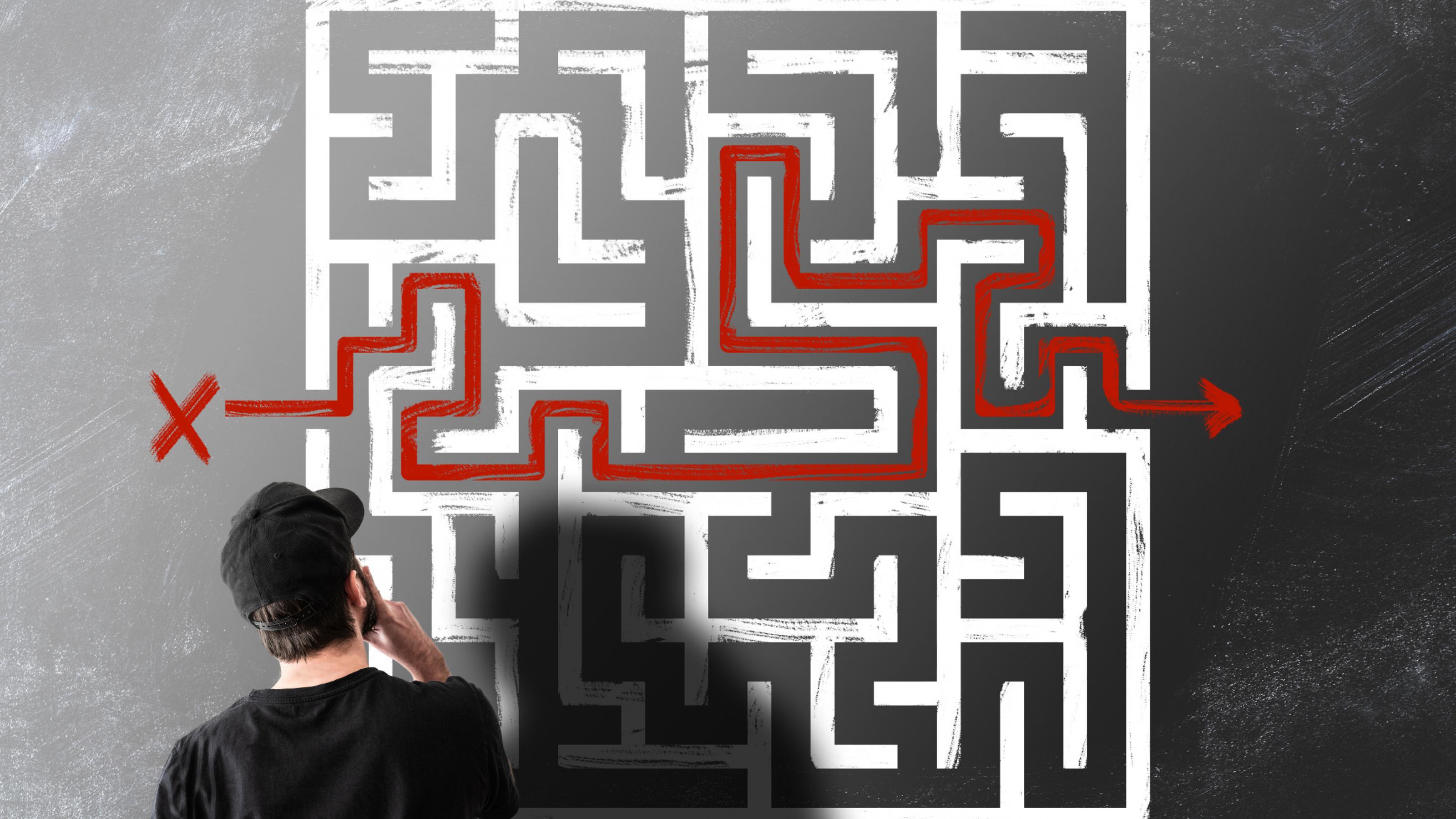 Mann mit Hut steht vor Bild eines Labyrinths, in das mit roter Farbe ein Weg eingezeichnet ist.