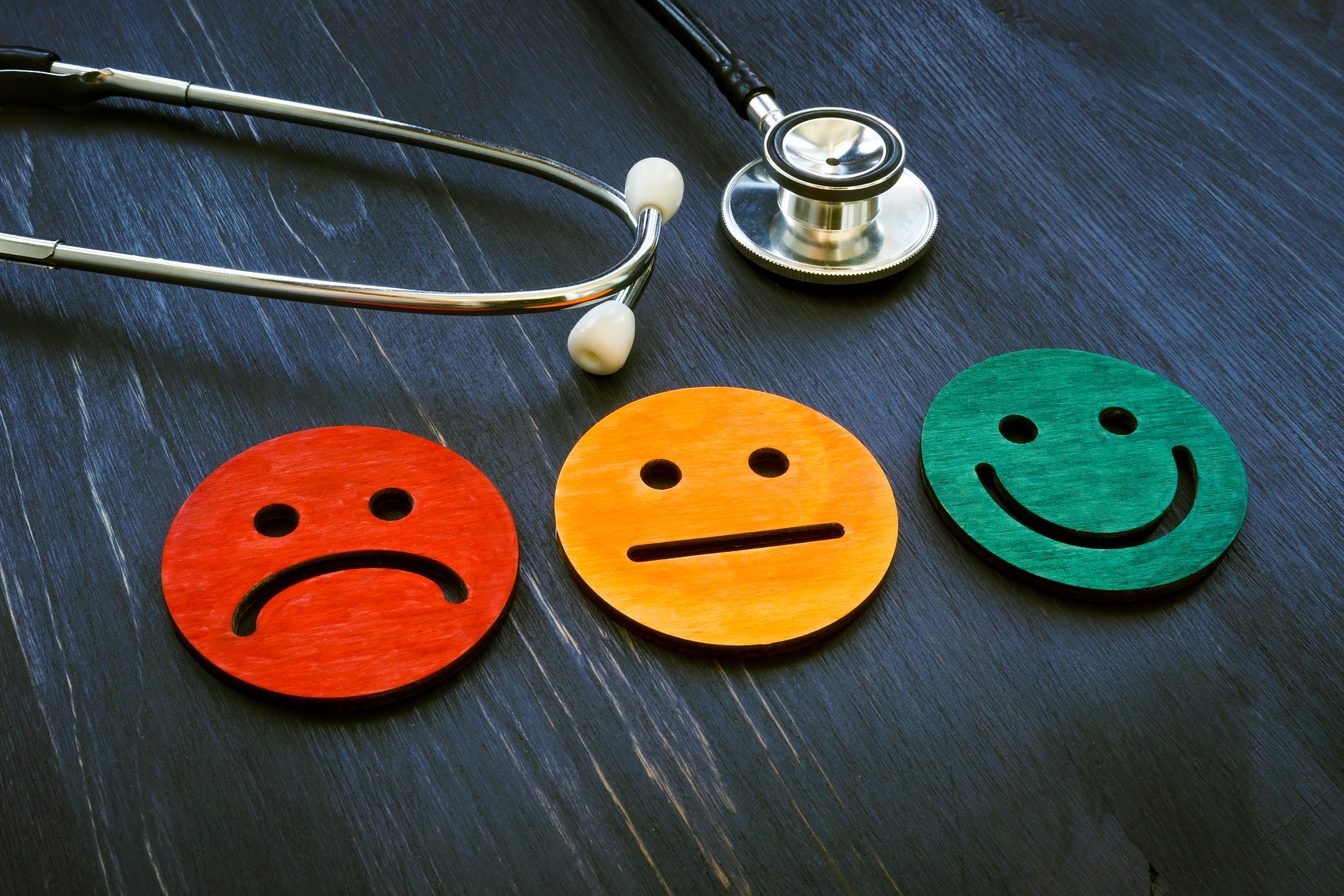 Ein Stethoskop liegt auf einem Tisch, darunter ein trauriger, ein neutraler und ein lachender Smiley in Rot, Gelb und Grün