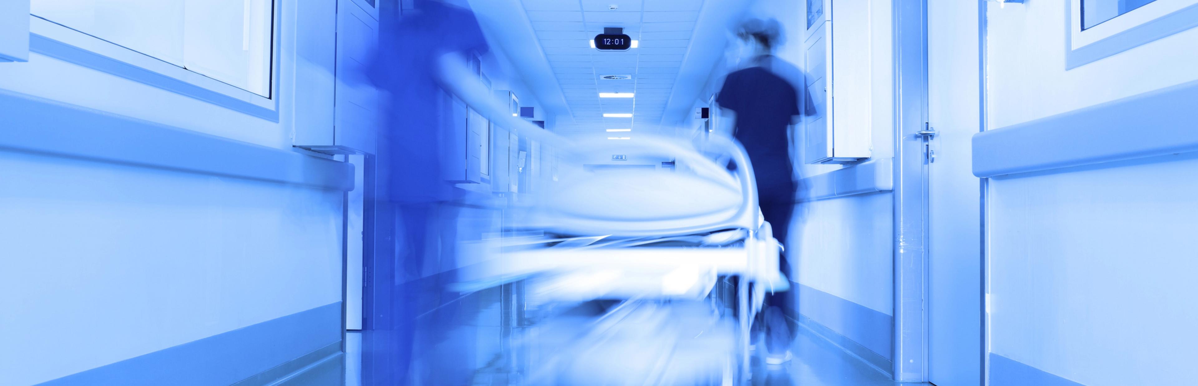 Zwei Pfleger:innen schieben ein Bett über einen Krankenhausflur. Foto zeigt die Szene verschwommen und mit blauem Filter