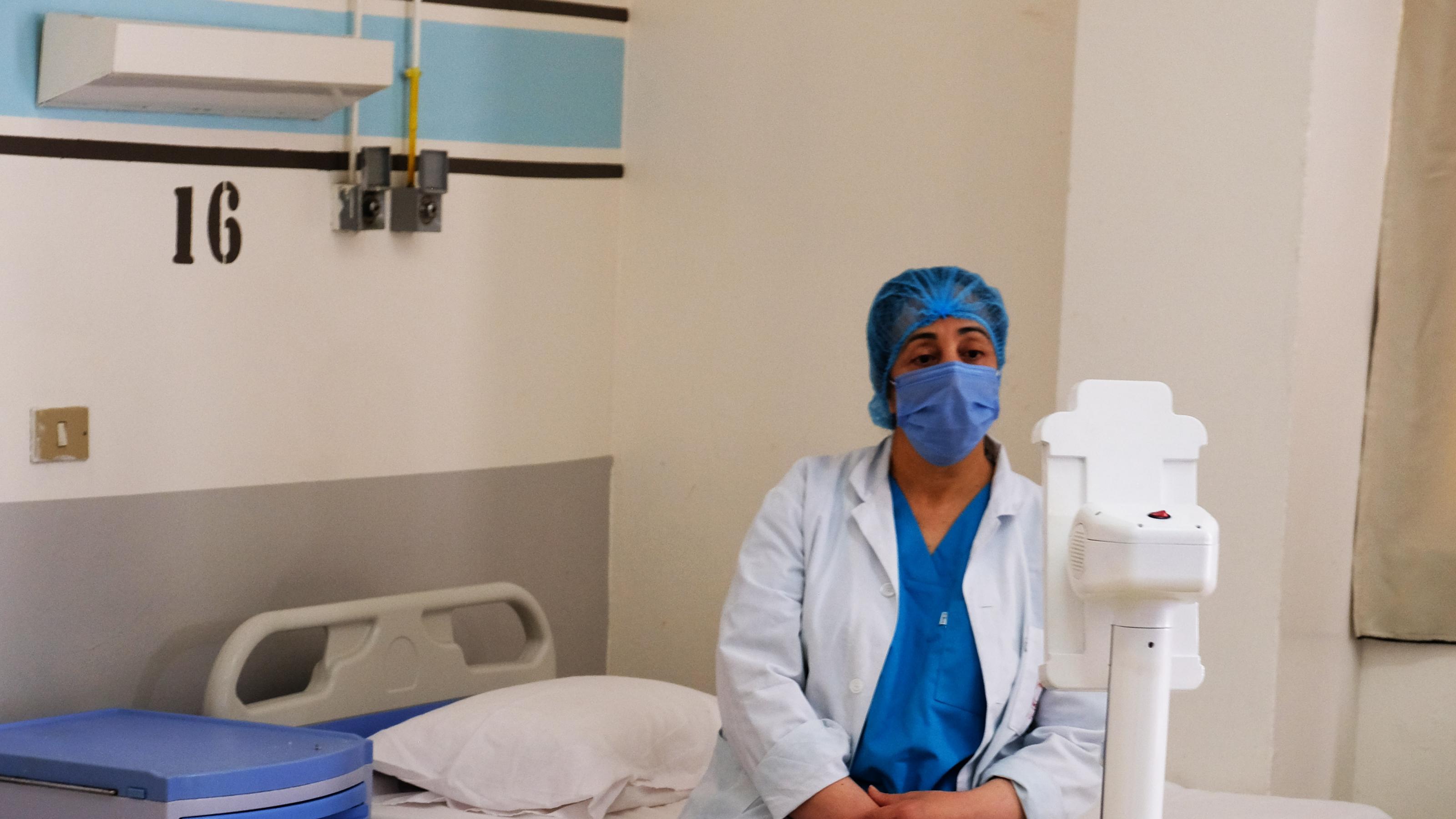 Eine Krankenpflegerin mit Kittel und Maske sitzt auf einem Krankenbett und schaut einen kleinen Roboter an.