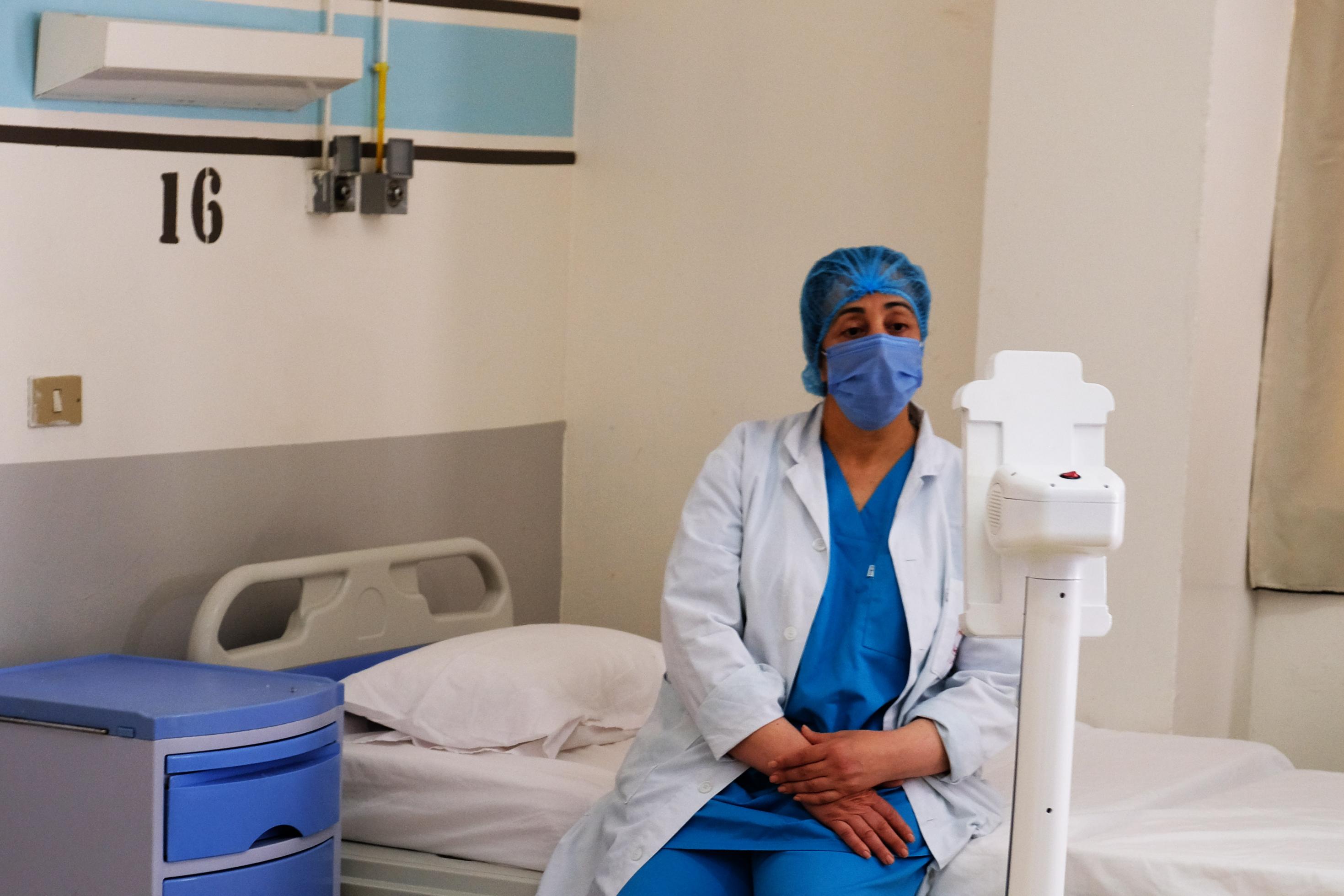 Eine Krankenpflegerin mit Kittel und Maske sitzt auf einem Krankenbett und schaut einen kleinen Roboter an.