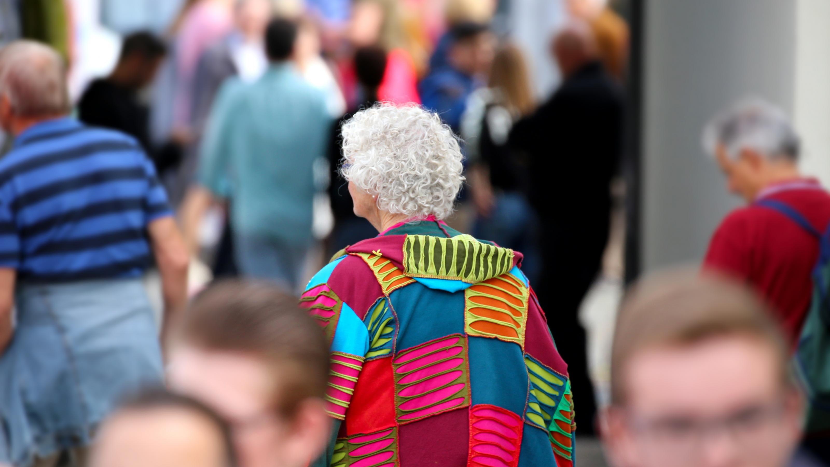 Menschen in einer Fußgängerzone. In der Mitte des Bildes eine ältere Frau von hinten, mit grauen Haaren und einer bunten Jacke.
