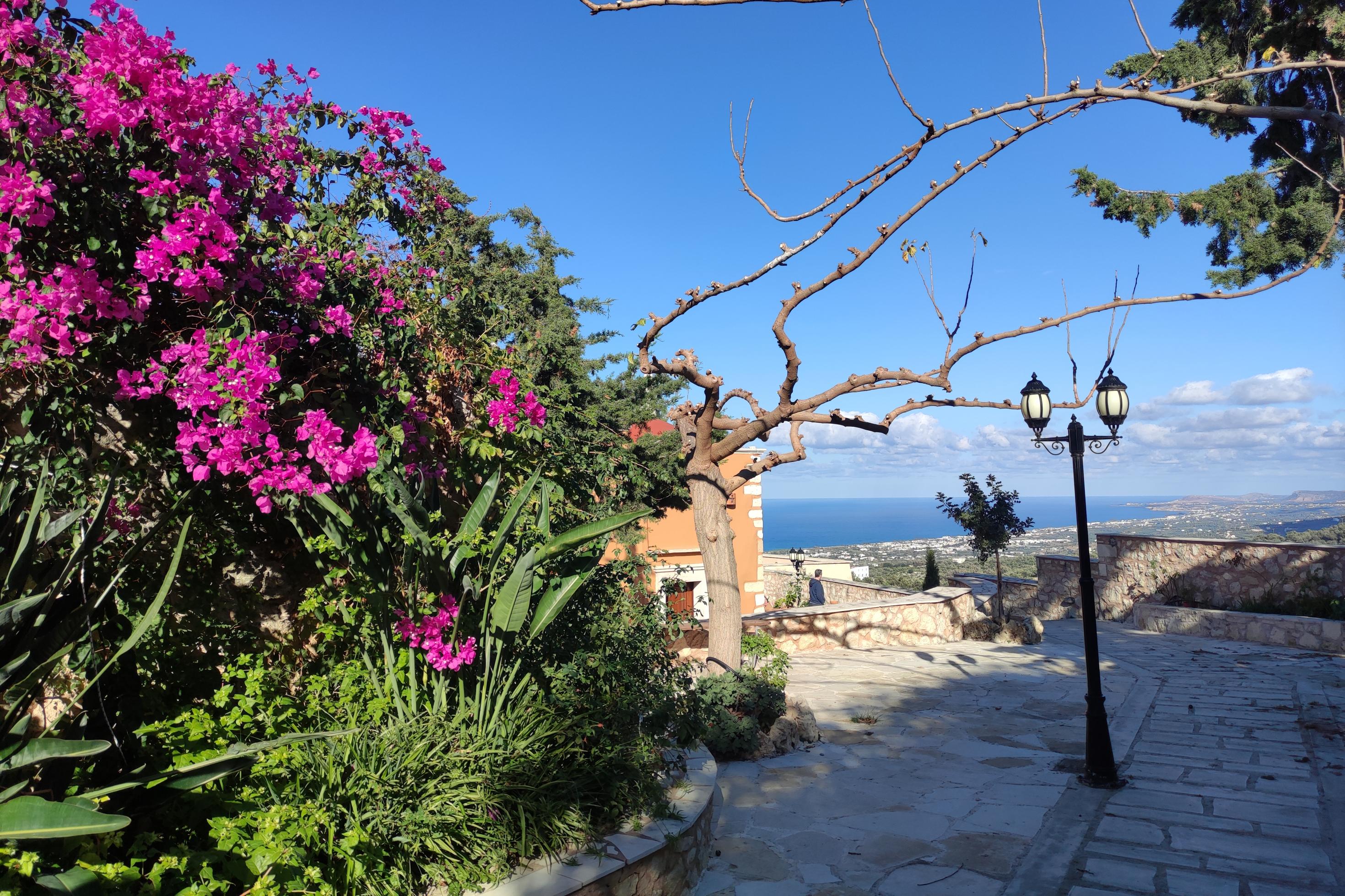 Ein Weg aus Steinplatten führt an einer üppigen Wand aus leuchtend pinken Bougainvillea-Blüten vorbei und öffnet den Blick auf das blaue Meer in der Ferne, das fast nahtlos in den blauen Himmel übergeht. Die Landschaft liegt unterhalb weit ausgebreitet.