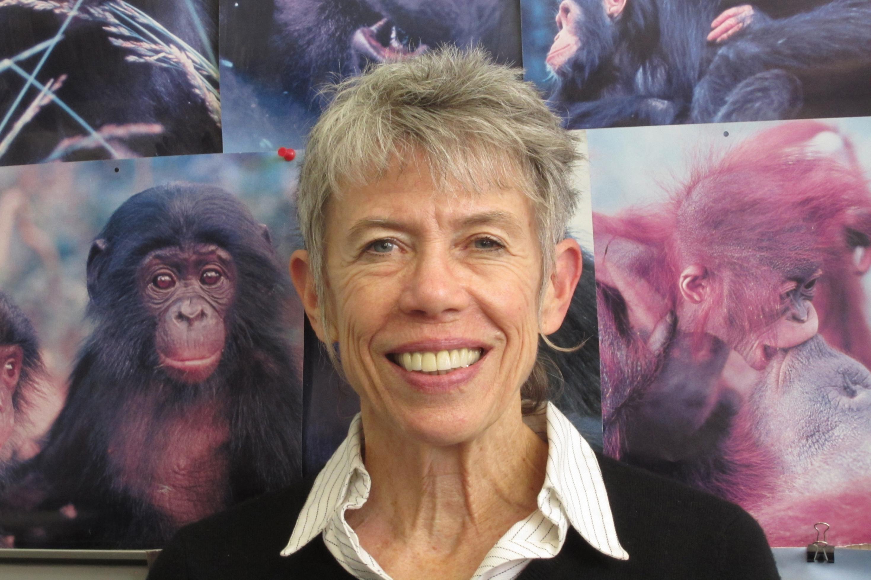 Das Gesicht einer Frau mit kurzen, grauen Haaren, vielen Lachfalten im Gesicht und zu einem Lächeln entblößten großen Zähnen ist zu sehen. Hinter ihr eine Wand mit Porträtfotos von Schimpansen, Bonobos, Organ-Utans und Gorillas.