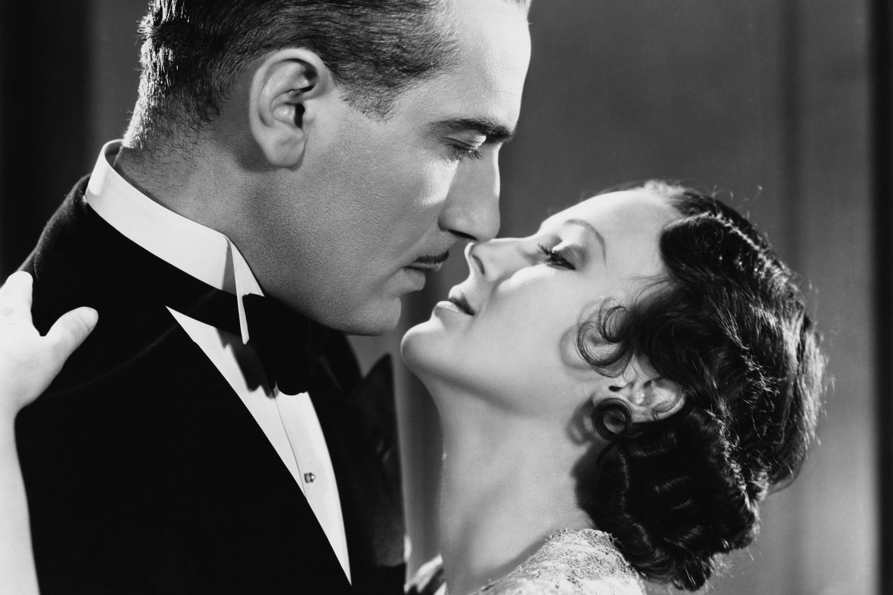 Eine Frau und ein Mann in eleganter Kleidung stehen eng zusammen und sind kurz davor sich zu küssen. Szene aus einem 1920er Jahre Film.