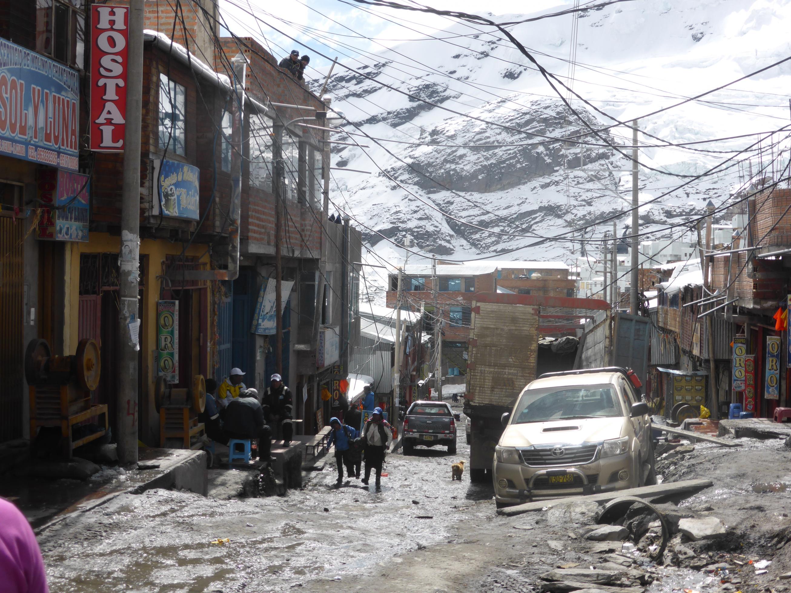 Abschüssige Strasse gesäumt von Geschäftshäusern, im Hintergrund schneebedecktes Gebirge, man sieht auf der Strasse Menschen in Anoraks und Bergbaukleidung
