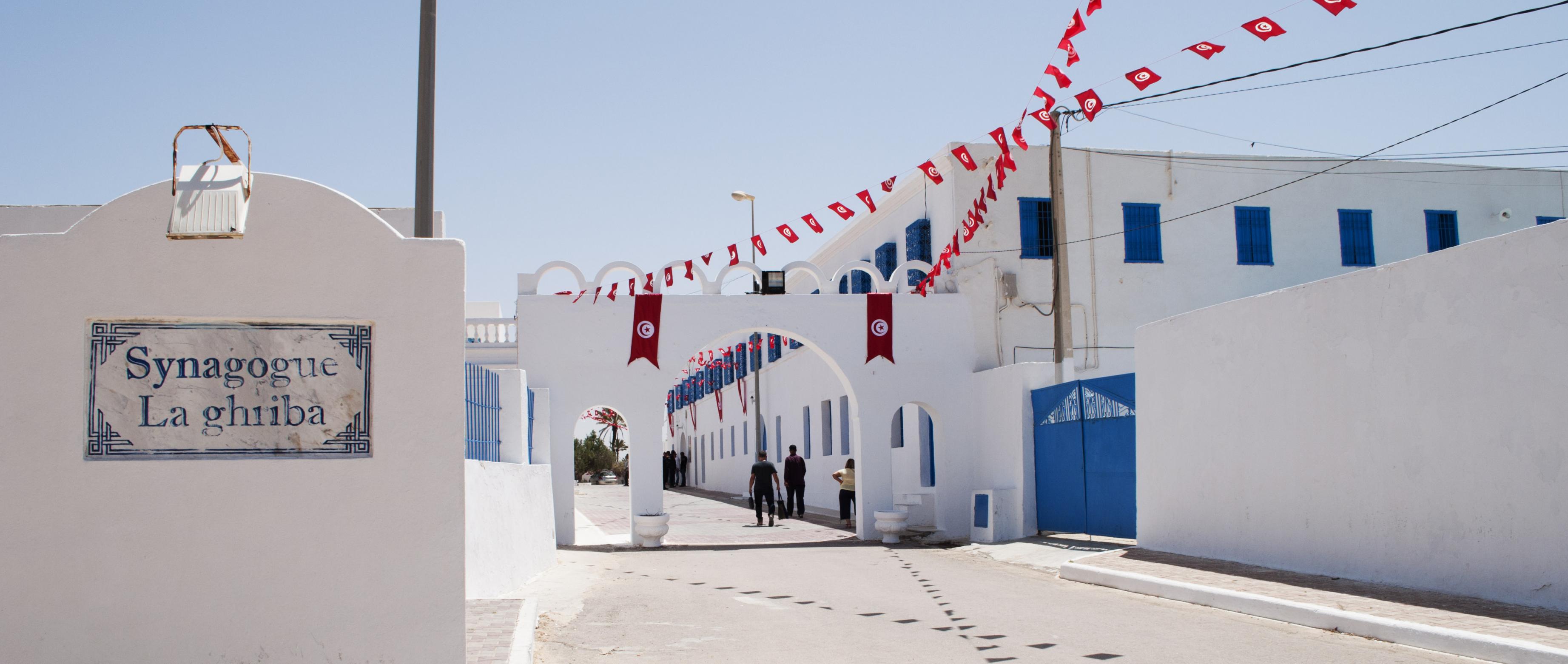 Einfahrt und Torbogen der Synagoge. Die Wände sind weiß getüncht, die Fenster blau vergittert, an den Dächern hängen tunesische Fahnen