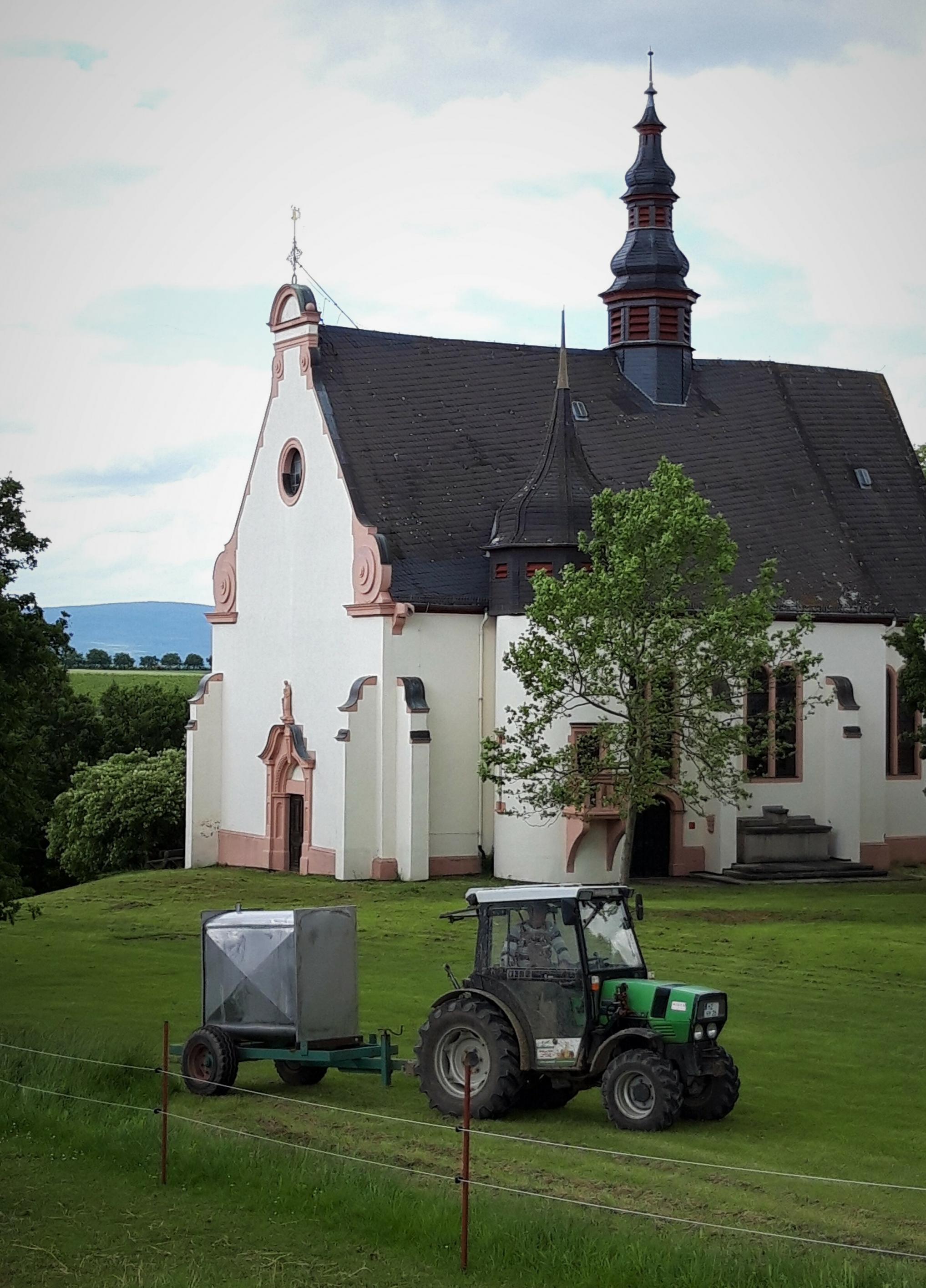 Eine kleine Barockkirche auf einer Anhöhe, umrahmt von einer Wiese, auf der ein kleiner Traktor unterwegs ist.