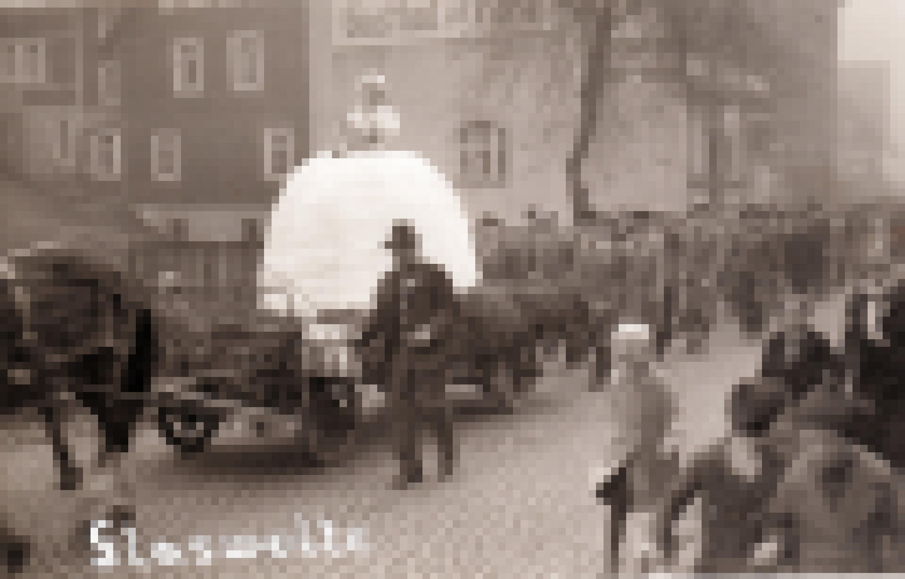 Ein altes Foto einer Parade. Auf einem Wagen der von Pferden gezogen wird ist eine Frau mit einem leuchtend weißen Kleid. Dahinter läuft eine Gruppe Männer in Anzügen.
