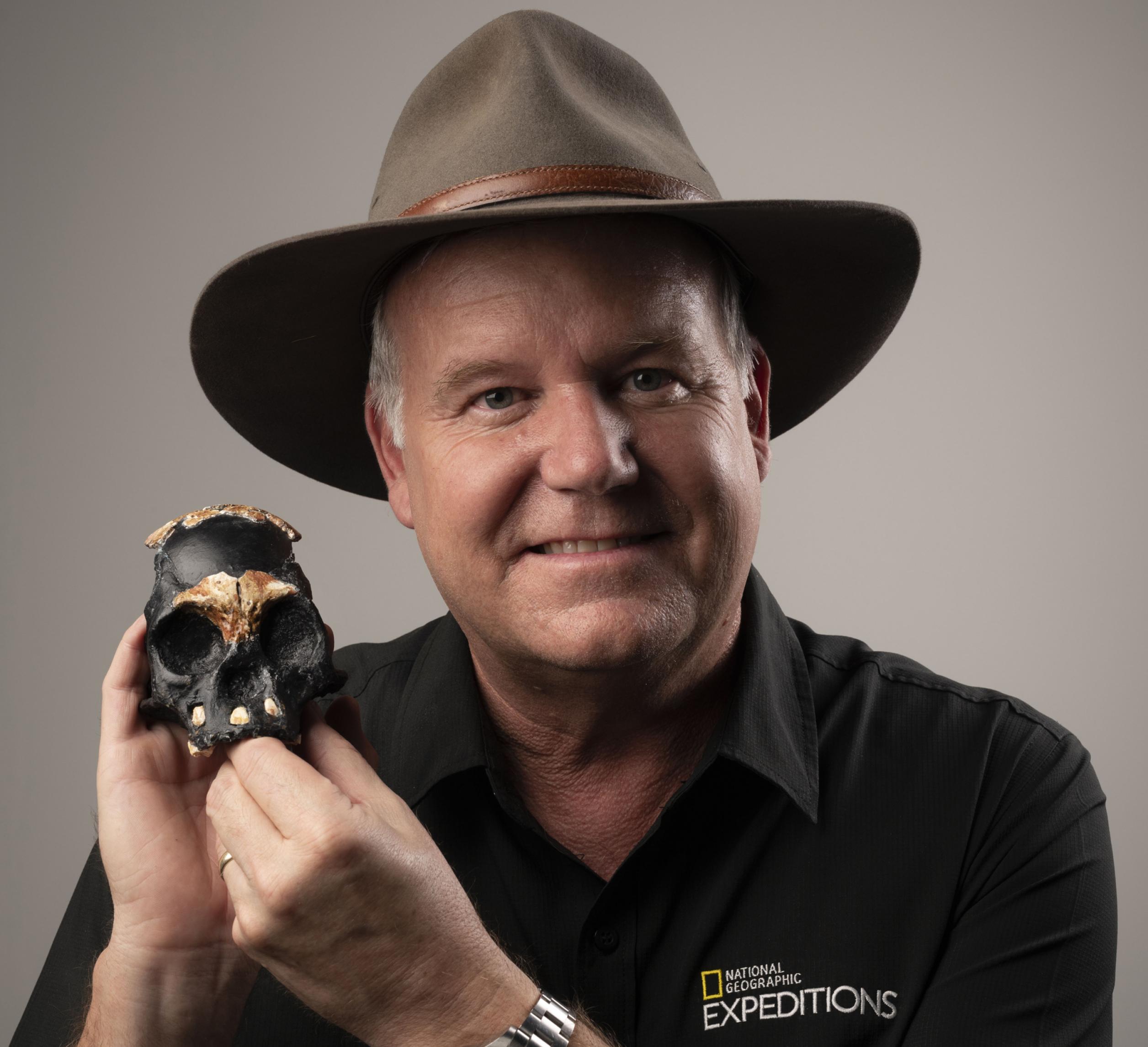 Das Foto zeigt einen Mann, Mitte fünfzig, mit braunem Filzhut und schwarzem Hemd, der in seiner rechten Hand einen etwa faustgroßen Schädel, zusammengesetzt aus hellbraunen fossilen Knochen und schwarzem Material, trägt.