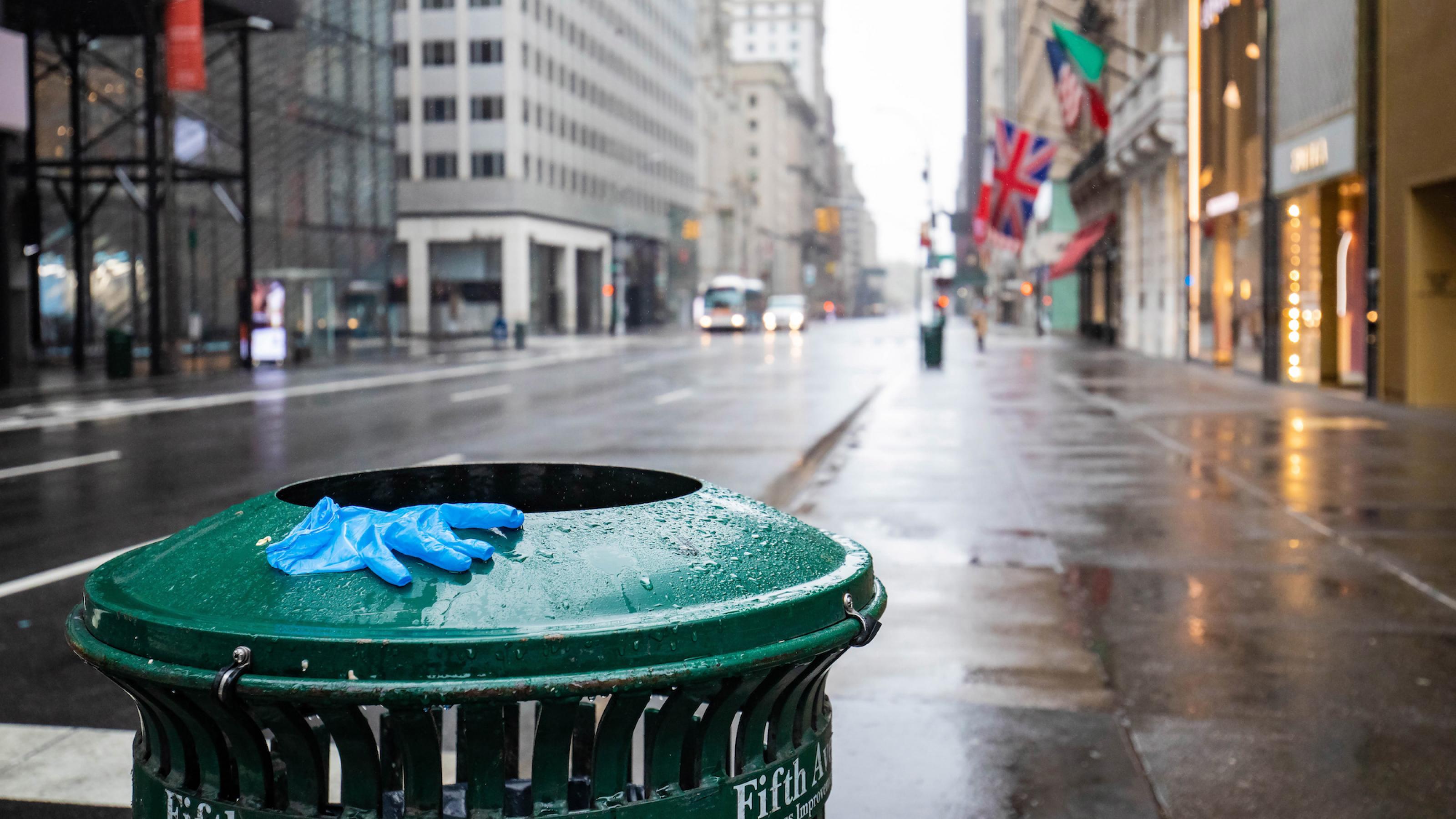 Das Bild zeigt einen Abfalleimer, auf dem ein Einweghandschuh liegt, dahinter eine leere Straße.