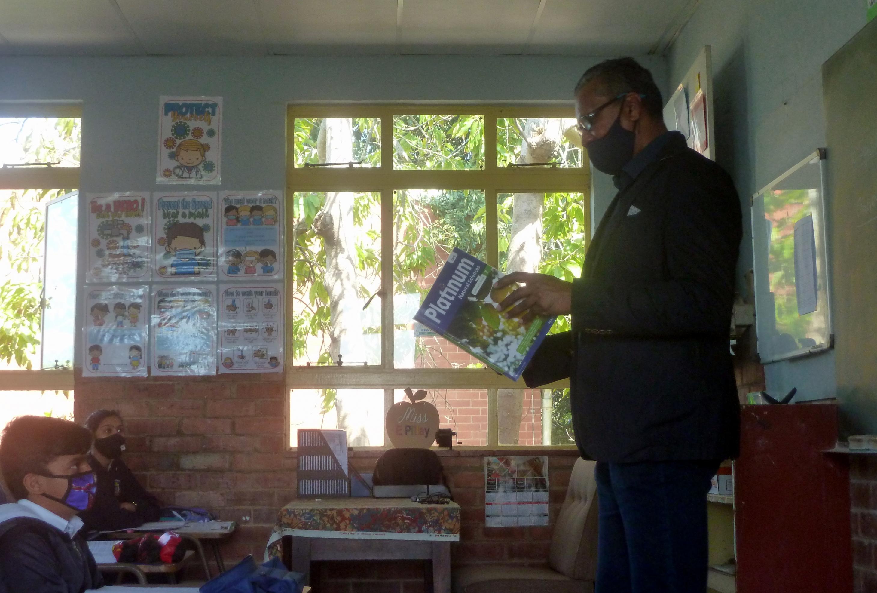 Der Lehrer steht mit einem Buch vor seiner Klasse, die Schüler schauen ihn an, an der Wand hängen Corona-Aufklärungsposter
