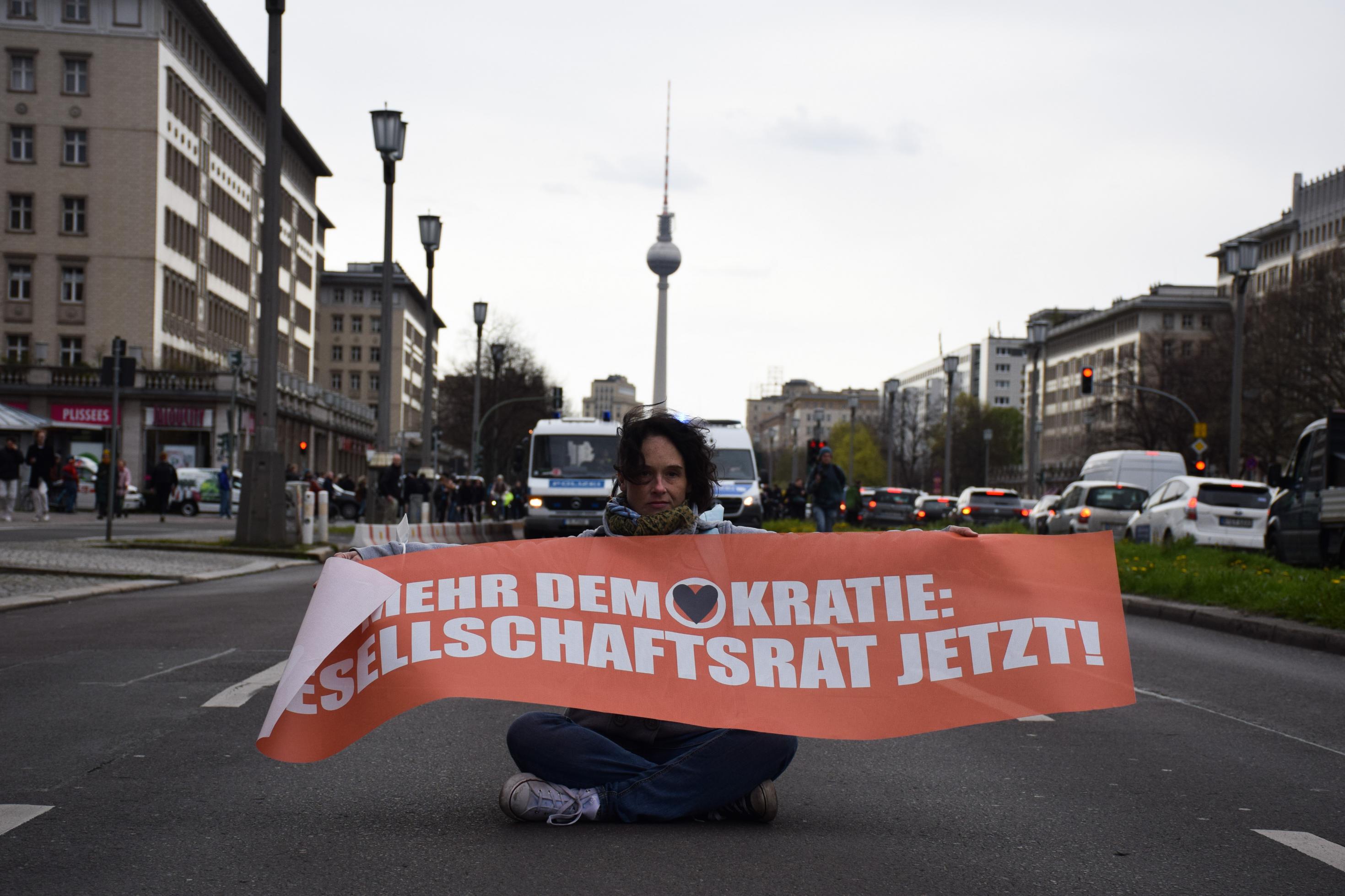 Eine Frau sitzt im Schneidersitz auf der Straße. Sie hält ein Transparent in der Hand mit der Aufschrift „Mehr Demokratie: Gesellschaftsrat jetzt!“. Im Hintergrund sieht man den Berliner Fernsehturm.