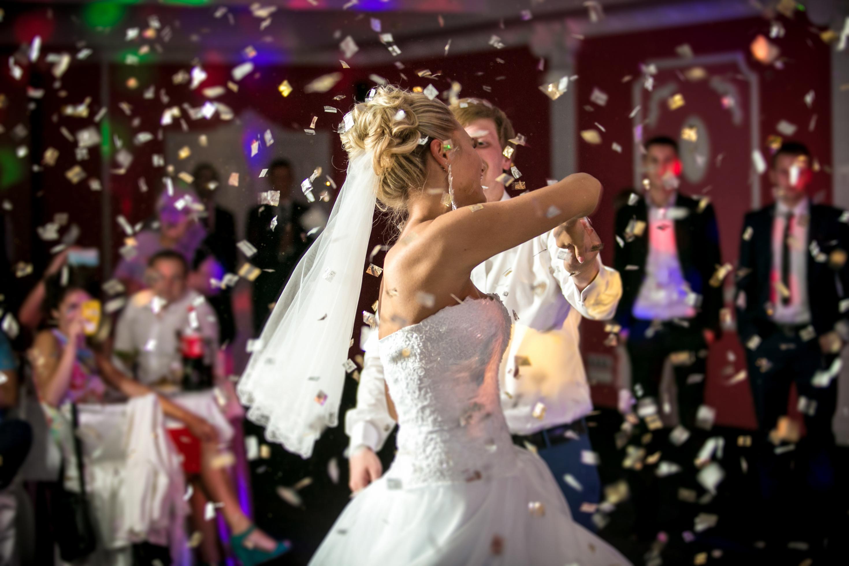 Eine Braut in weißem Kleid – Schulter und Oberarme frei, an den hochgesteckten blonden Haaren einen Schleier tragend – tanzt beschwingt mit ihrem Bräutigam, der großenteils von ihr verdeckt wird. Im Hintergrund unscharf und dunkel die zuschauenden Hochzeitsgäste und um das Paar herum ein von oben kommender Konfetti-Regen.