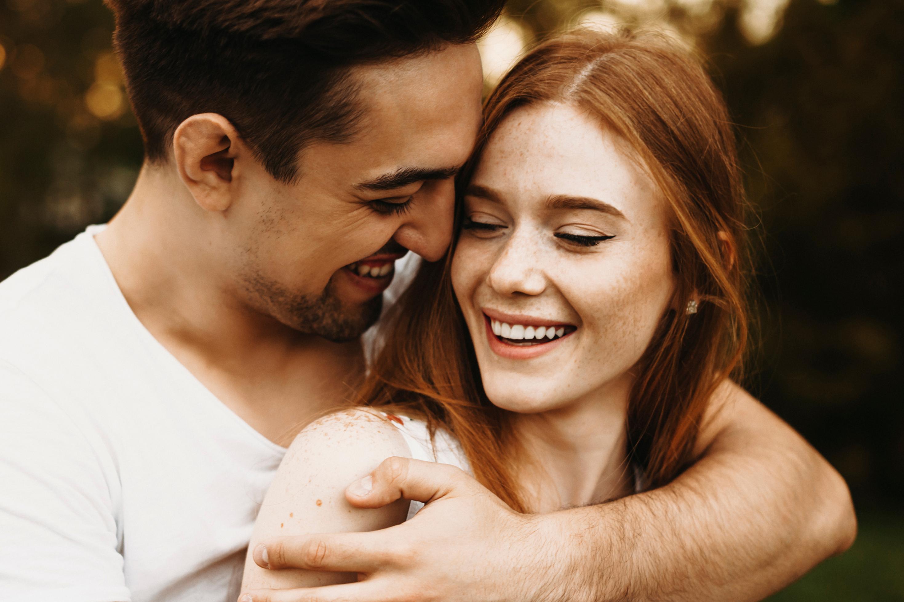 Zu sehen ist die Nahaufnahme eines Paares. Der Mann im weißen T-Shirt hat seine Partnerin von hinten mit dem Arm umfasst, ihr das Gesicht zugeneigt. Sie – im ärmellosen Sommerkleid und mit langen, glatten braunen Haaren – lächelt glücklich, die Augen halb geschlossen.