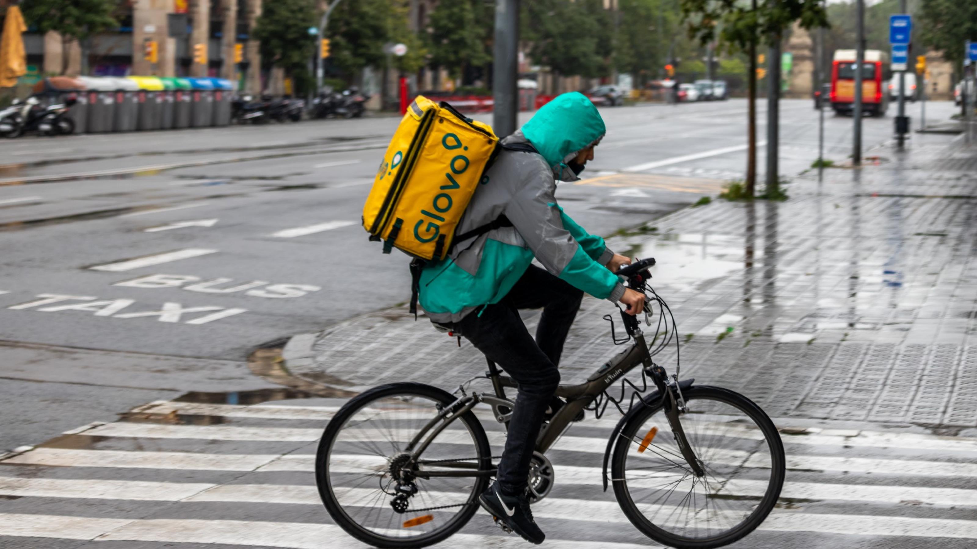 Ein Lieferbote fährt in einer spanischen Stadt über eine regennasse Straße, auf dem Rücken eine der charakteristischen quadratischen Lieferboxen.