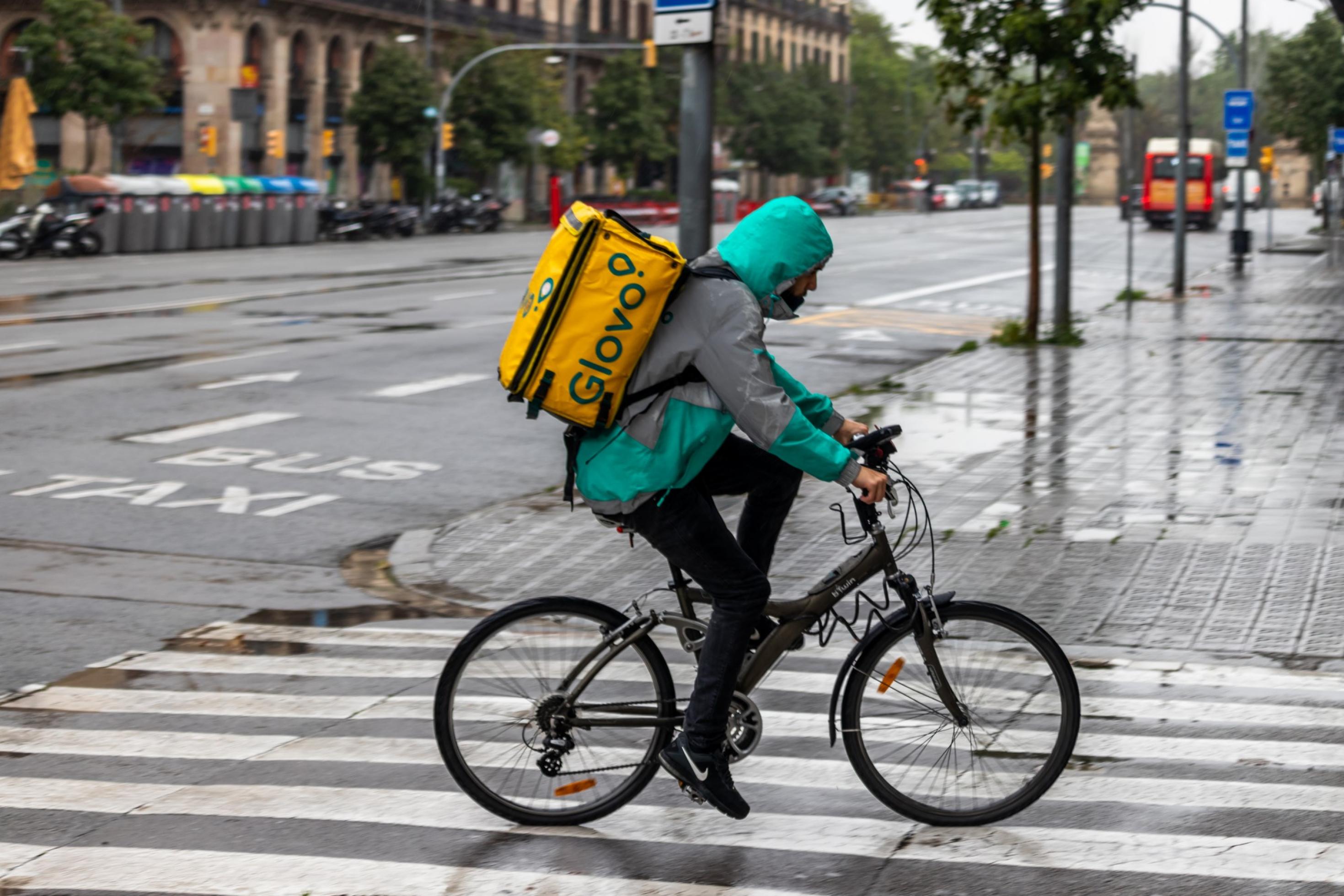 Ein Lieferbote fährt in einer spanischen Stadt über eine regennasse Straße, auf dem Rücken eine der charakteristischen quadratischen Lieferboxen.