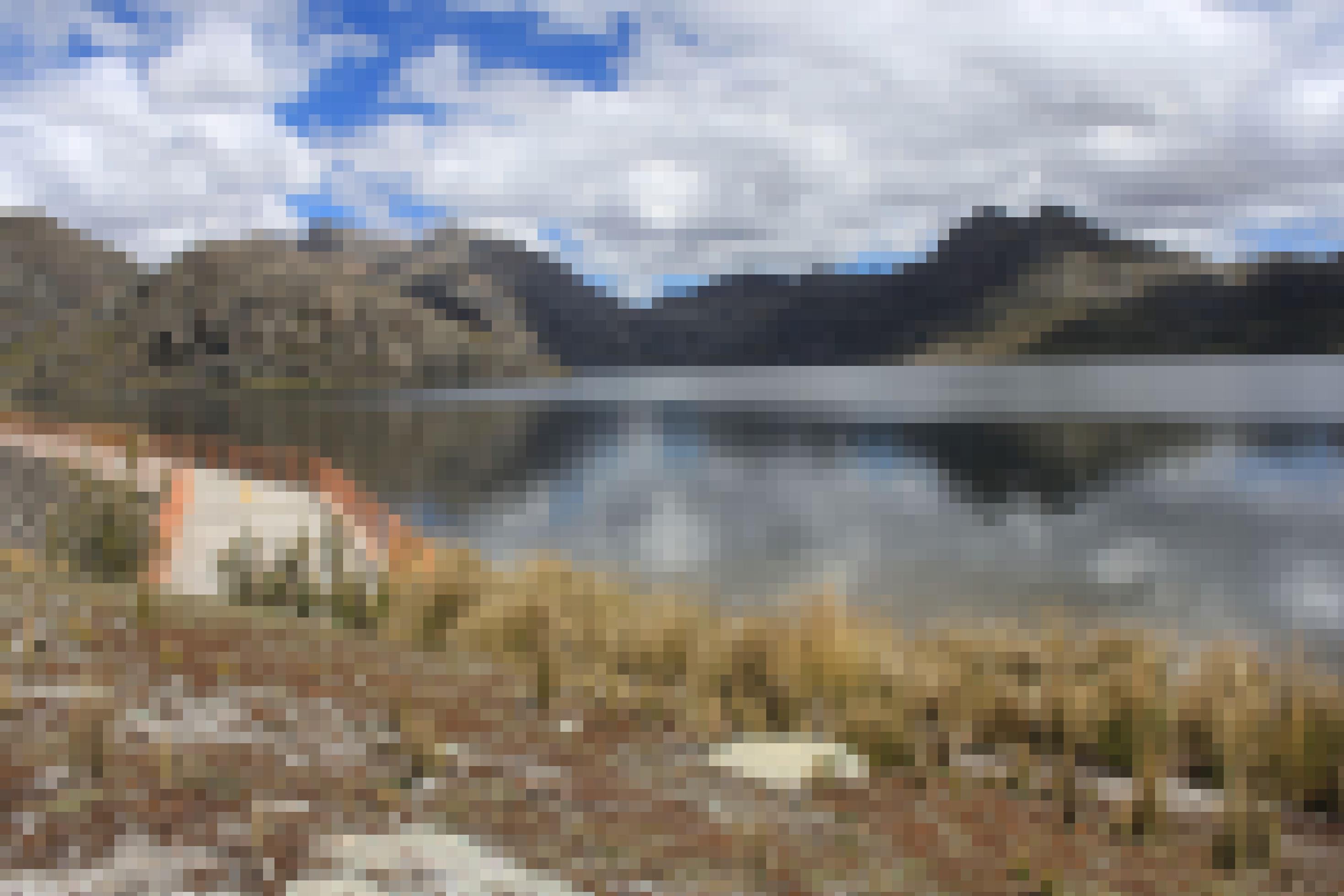 Berglandschaft mit Lagune. An der Lagune führt ein betonnierter Steg mit Gitter entlang für Arbeiten am Stausee