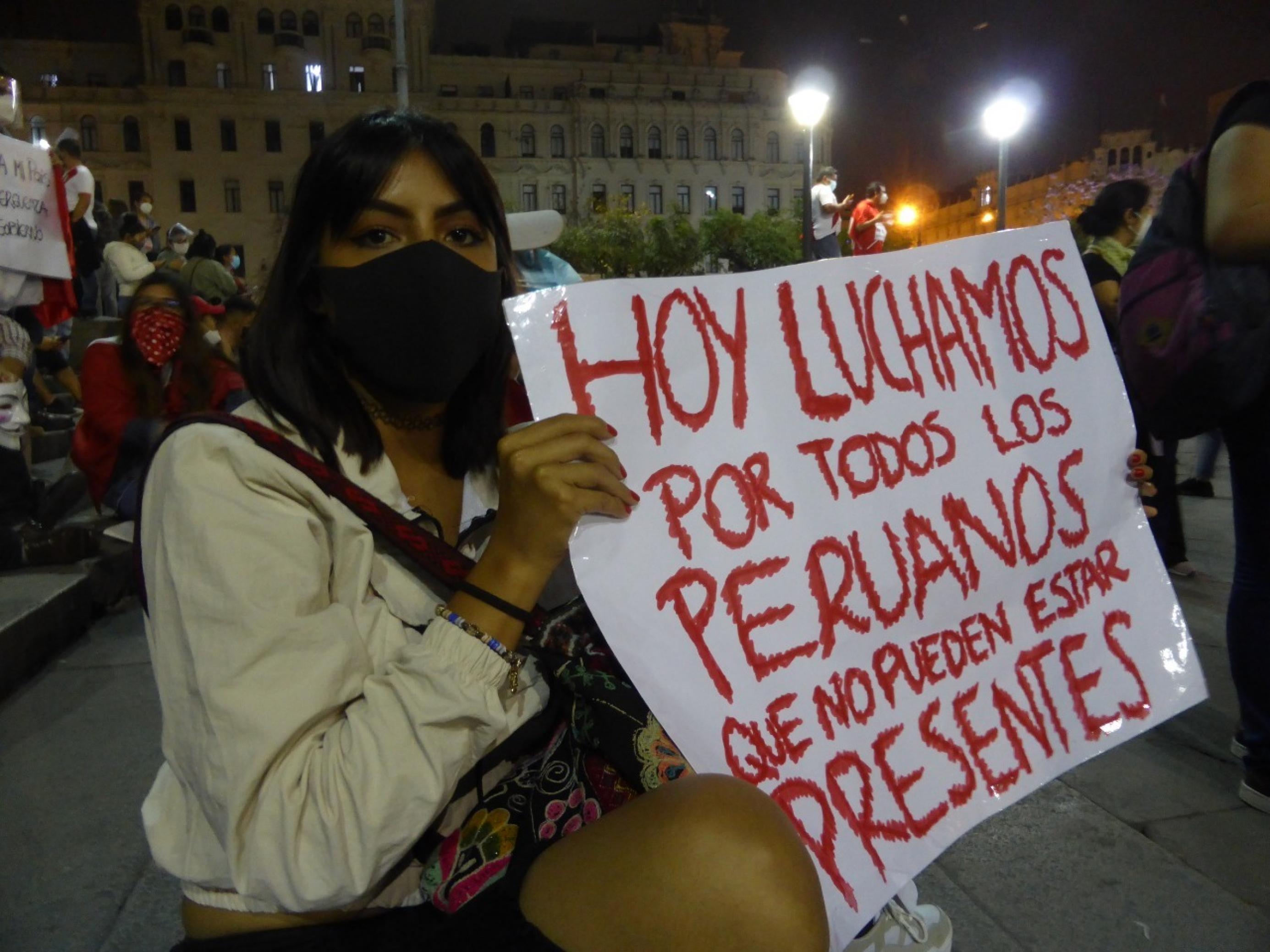 Junge Frau sitzt auf den Stufen eines grossen historischen Platzes in Lima. Neben sich ein Plakat. „Hoy luchamos por todos los peruanos que no pueden estar presentes“. „Heute kämpfen wir für all jene Peruaner, die nicht hier sein können“