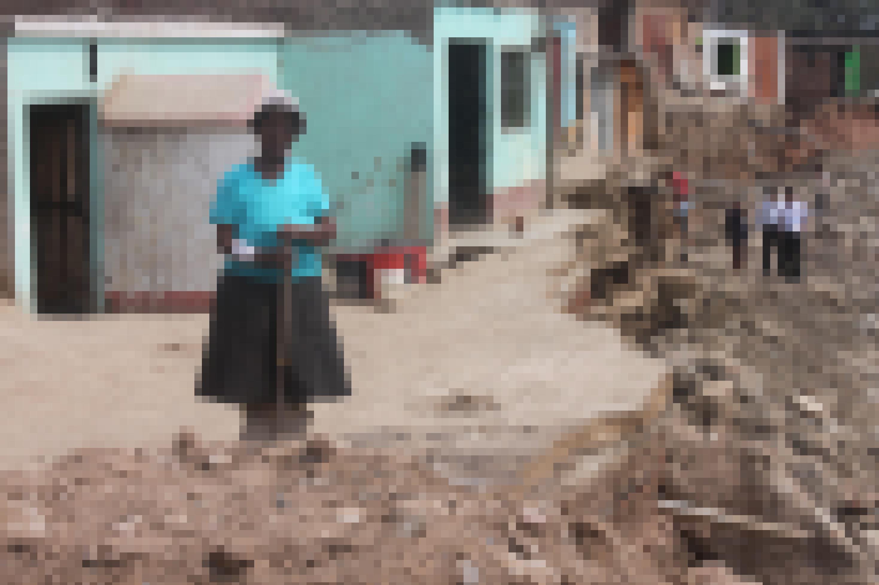 Ältere Frau mit türkisfarbenem T-Shirt, Hut und schwarzem knielangem Rock, hält eine Schaufel in der Hand. Im Hintergrund eine Häuserzeile. Die Strasse davor ist bis zur Tür einiger Häuser abgerissen.