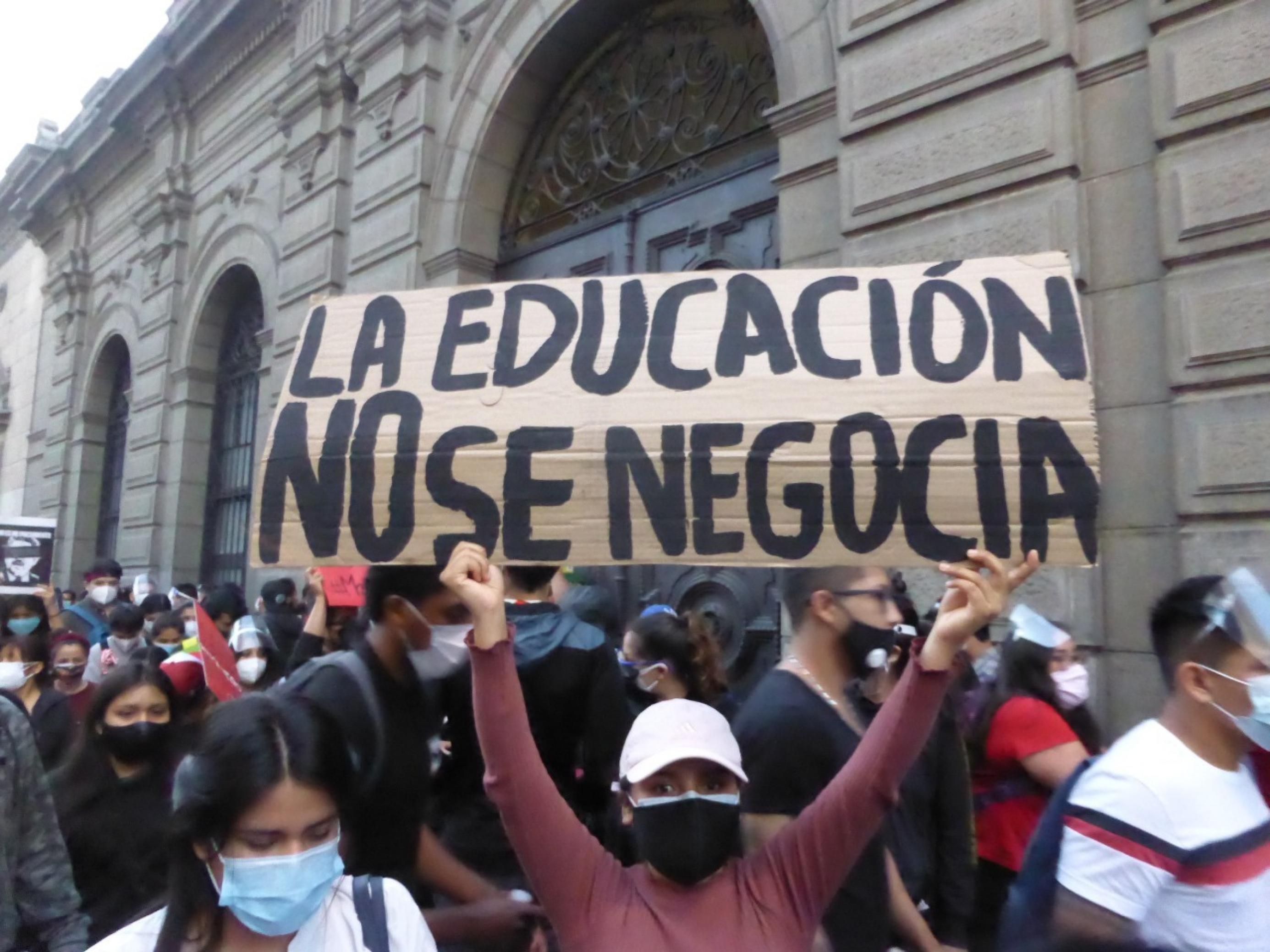 Mehrere demonstrierende Personen stehen zusammen. Eine Frau hält einen Karton hoch, auf dem aus Spanische steht: „Mit der Bildung macht man keine Geschäfte.“