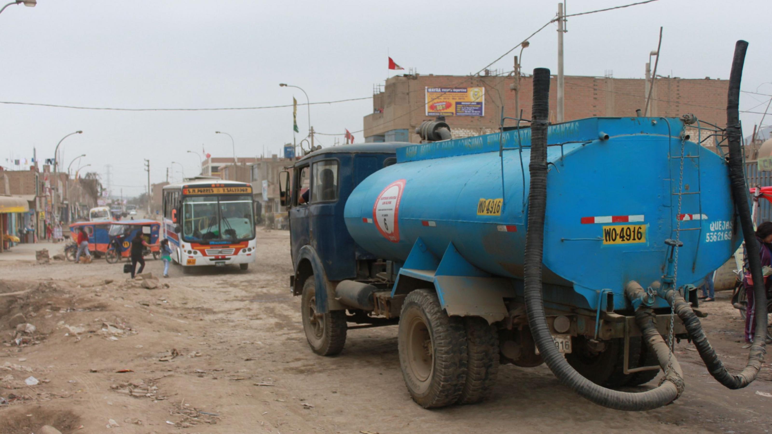 Ein blauer Tanklastwagen in einem Armenviertel, graue Erdstrasse, ein Bus, graue Häuser, kein Grün.