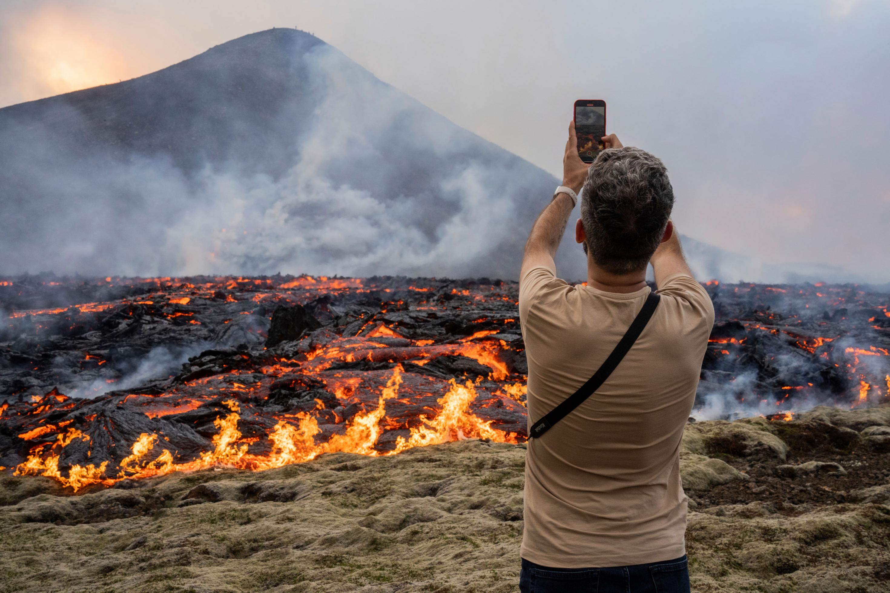 Ein Mann mit dem Rücken zum Betrachter steht vor einem Lavafeld, in dem es teilweise noch rot glüht und dampft. Er hält sein Telefon in die Höhe und macht ein Foto.