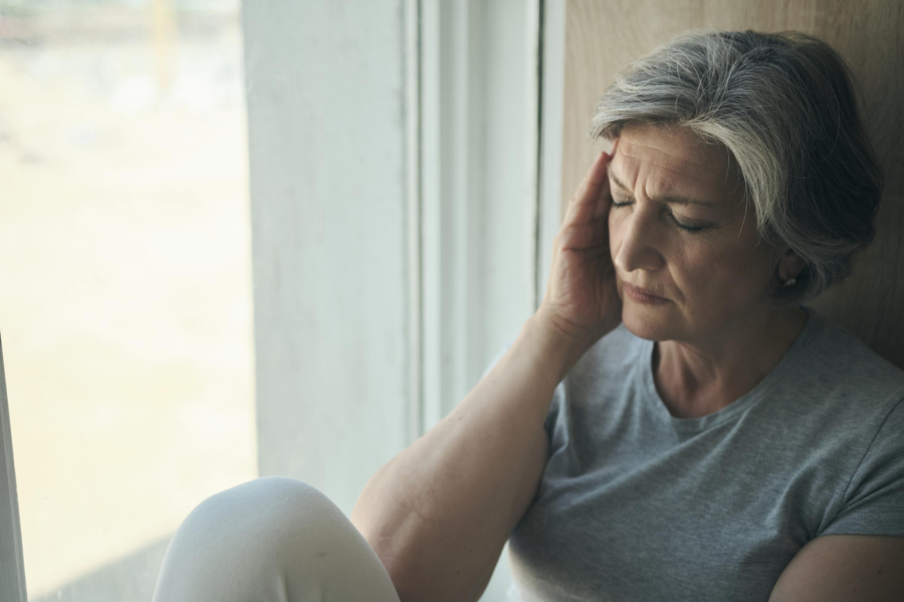 Eine ältere, erschöpfte Frau sitzt am Fenster und massiert ihre Schläfe wegen ihrer Kopfschmerzen, ein häufiges Symptom von Long Covid.pfsc