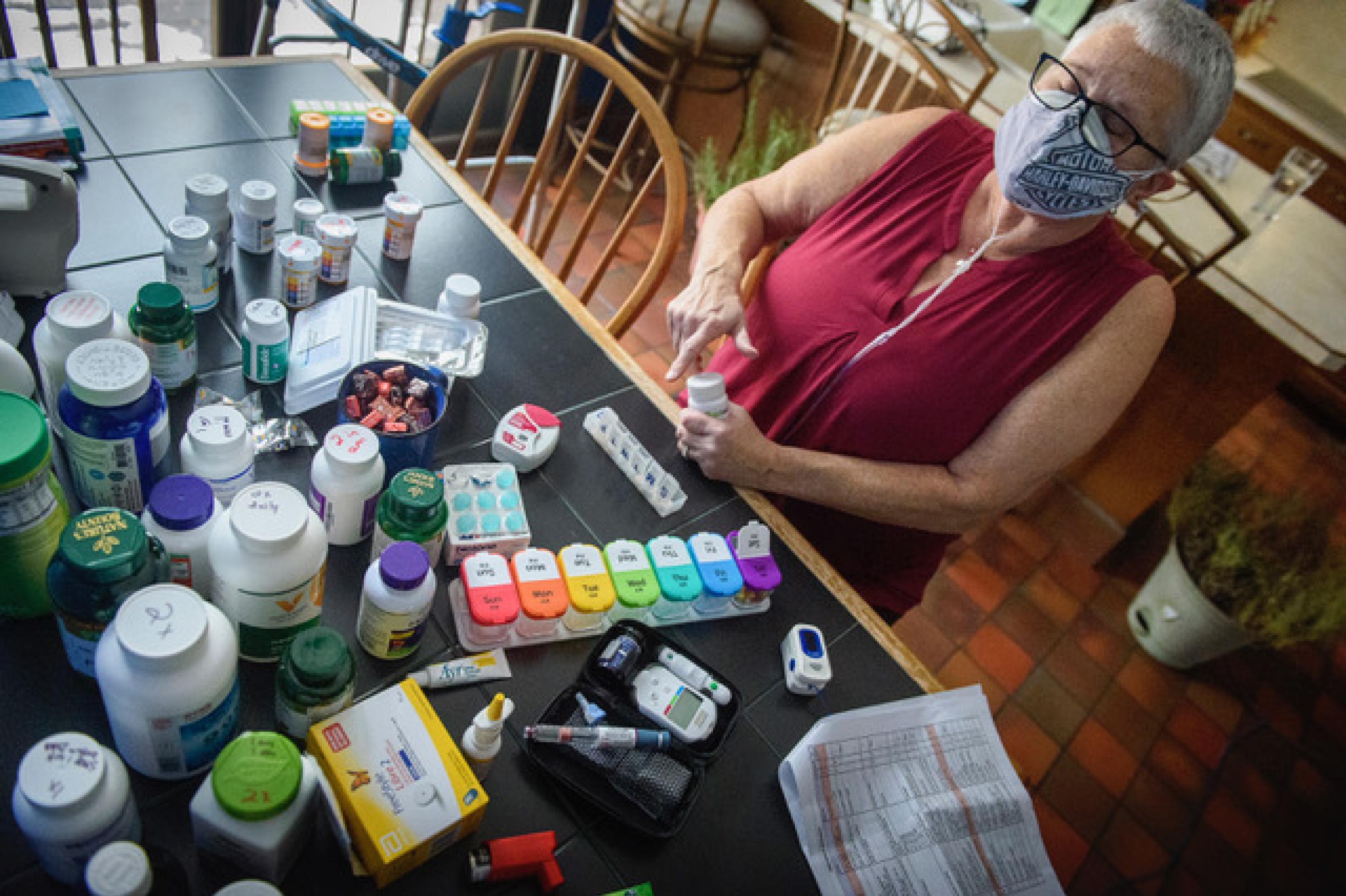 Eine Frau sitzt an einem Tisch, auf dem dutzende Medikamentenpackungen ausgebreitet sind. Die Frau trägt eine Maske, darunter ist ein Beatmungsschlauch zu sehen.