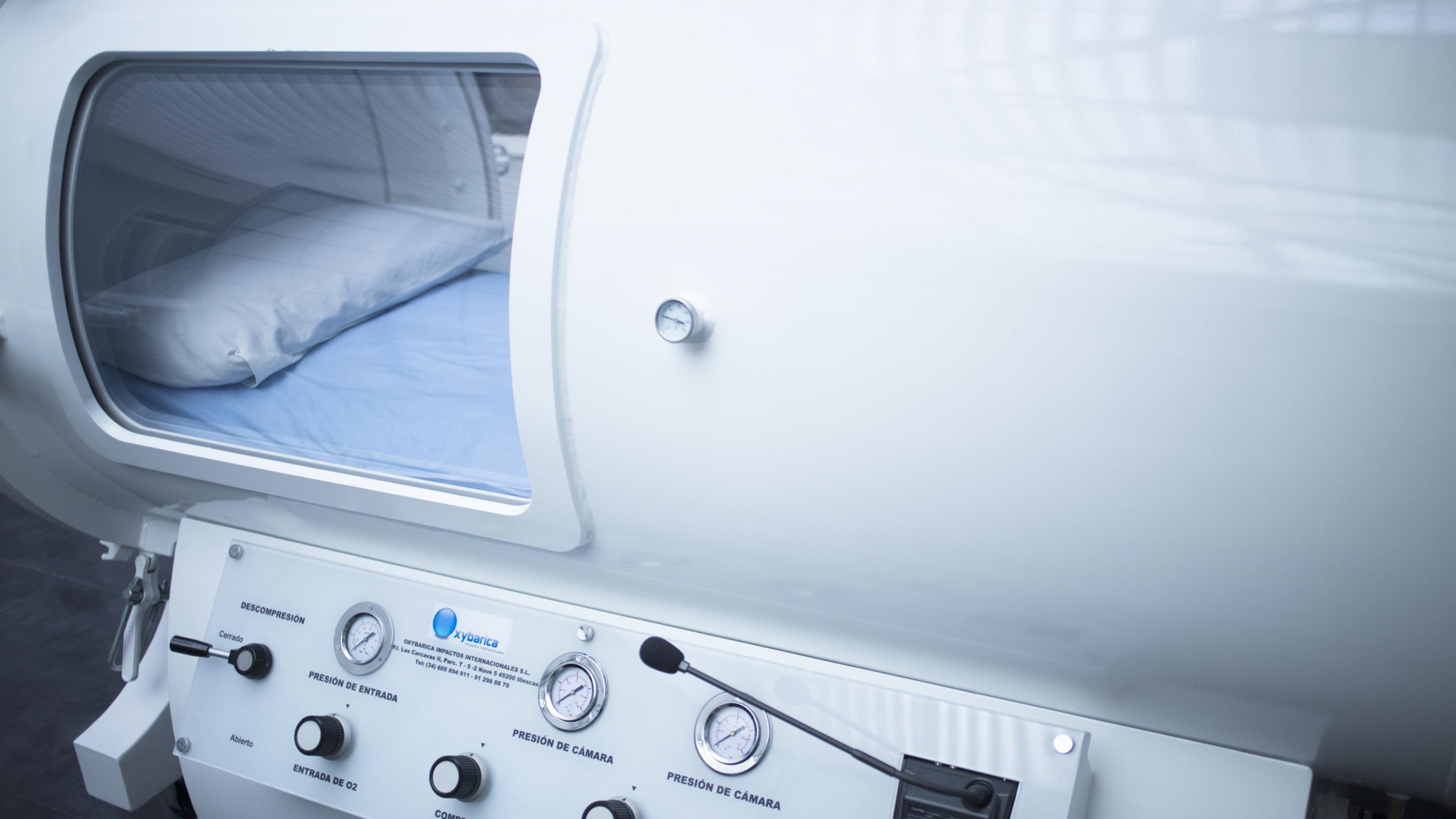 Eine Sauerstoff-Druckkammer in Form einer großen, weißen Kapsel mit Instrumenten zur Bedienung. Ein Fenster offenbart den Blick ins Innere der Kammer: Patient:innen können sich zur Behandlung hier auf eine Matratze und ein Kopfkissen legen.