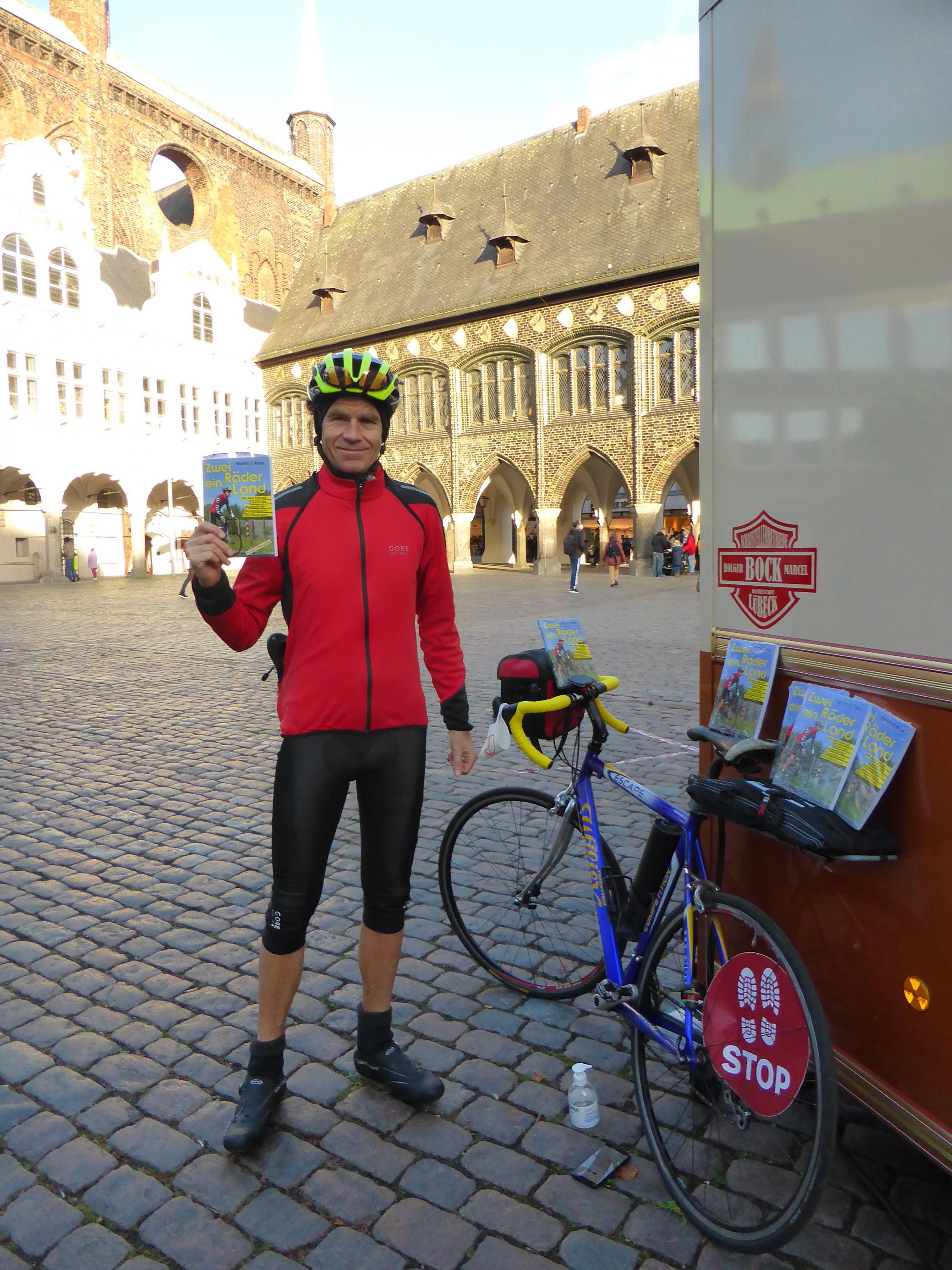 Martin C Roos steht in Radlerkluft neben seinem Rennrad, in der Rechten ein Exemplar seines Buchs „Zwei Räder, ein Land“.