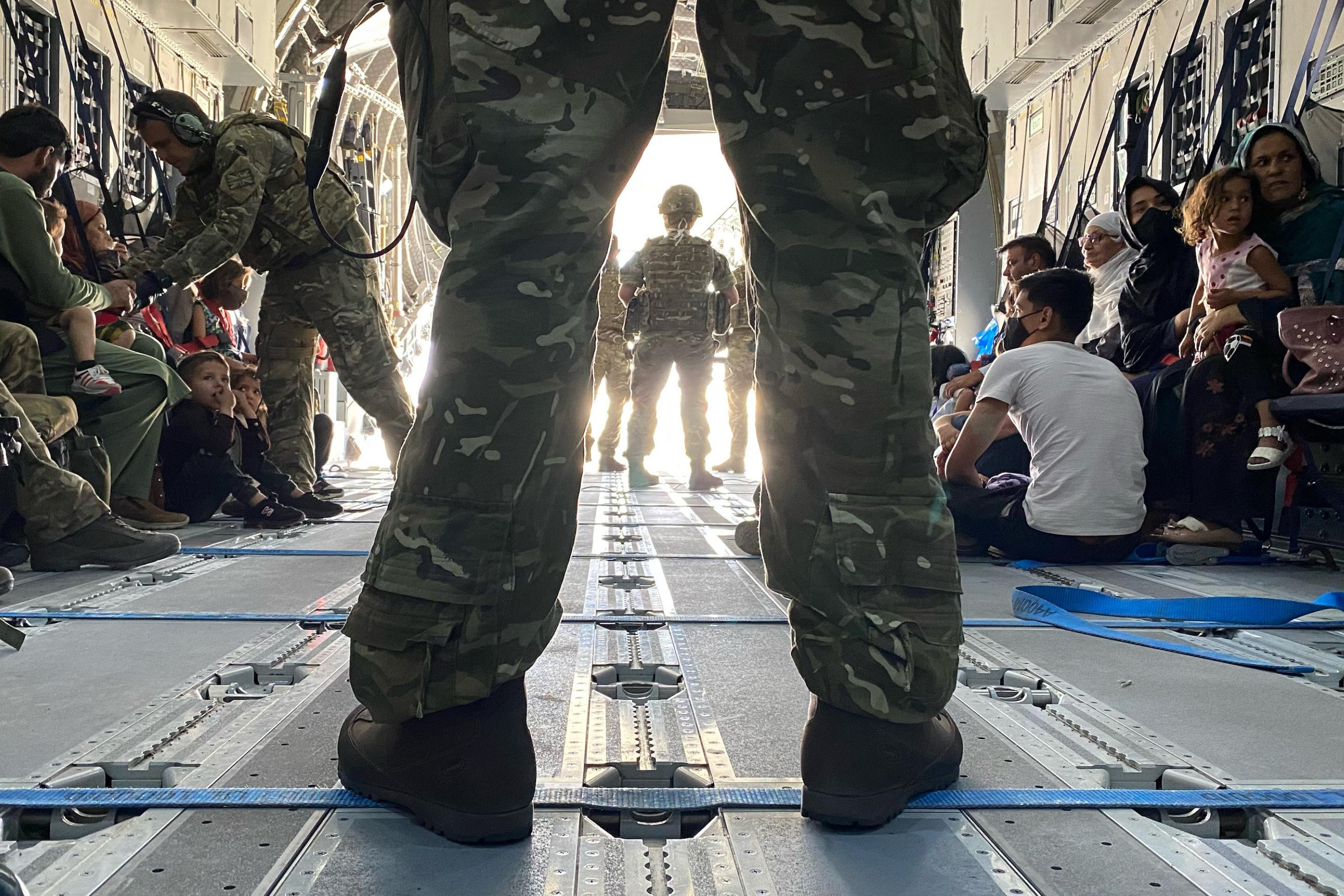 Blick in den Laderaum einer britischen Militärmaschine, im Vordergrund die Beine eines breitbeinig stehenden Soldaten in Tarnuniform. Im Hintergrund zivile Personen, die auf dem Boden des Flugzeuges sitzen.