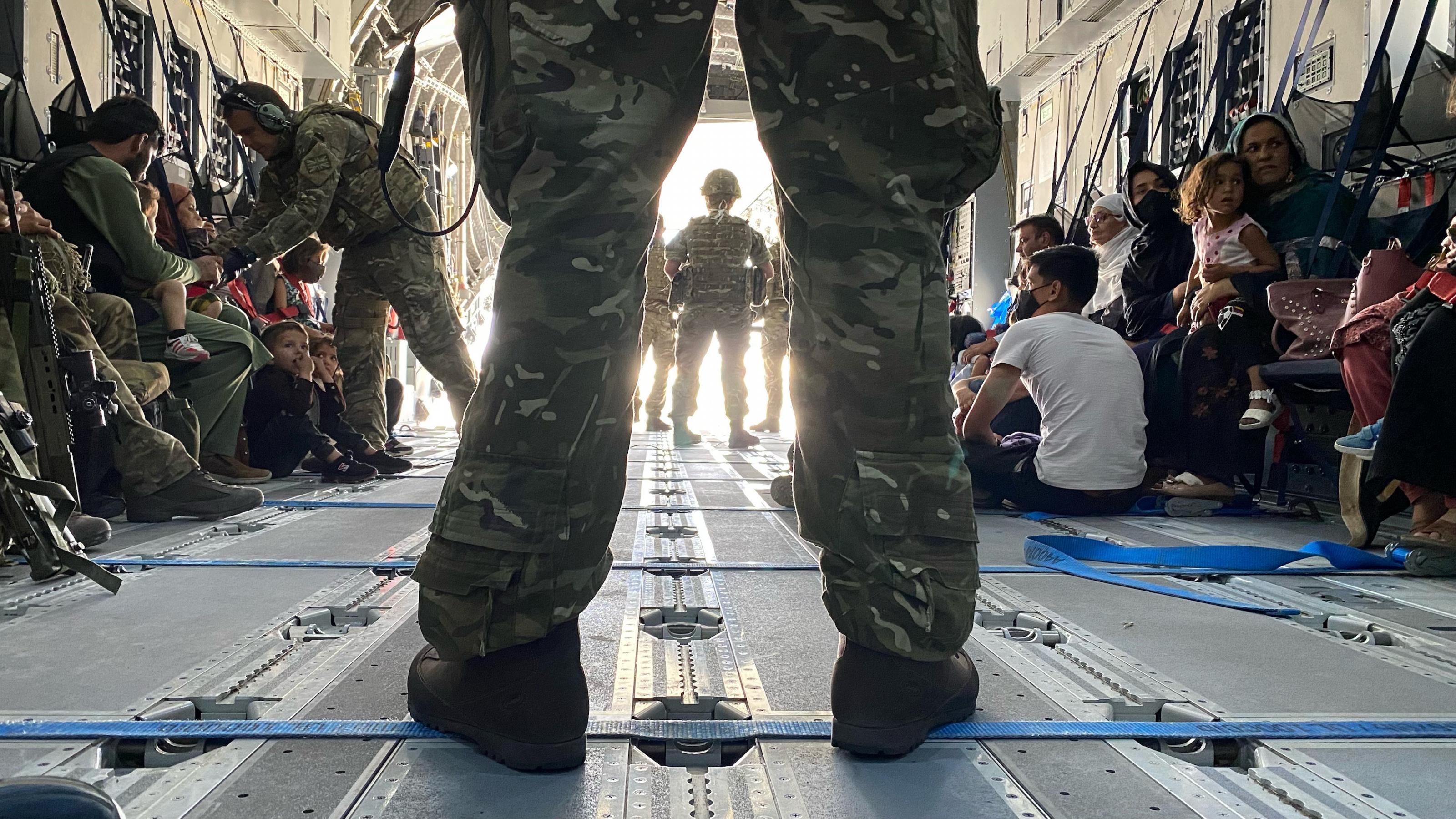 Blick in den Laderaum einer britischen Militärmaschine, im Vordergrund die Beine eines breitbeinig stehenden Soldaten in Tarnuniform. Im Hintergrund zivile Personen, die auf dem Boden des Flugzeuges sitzen.