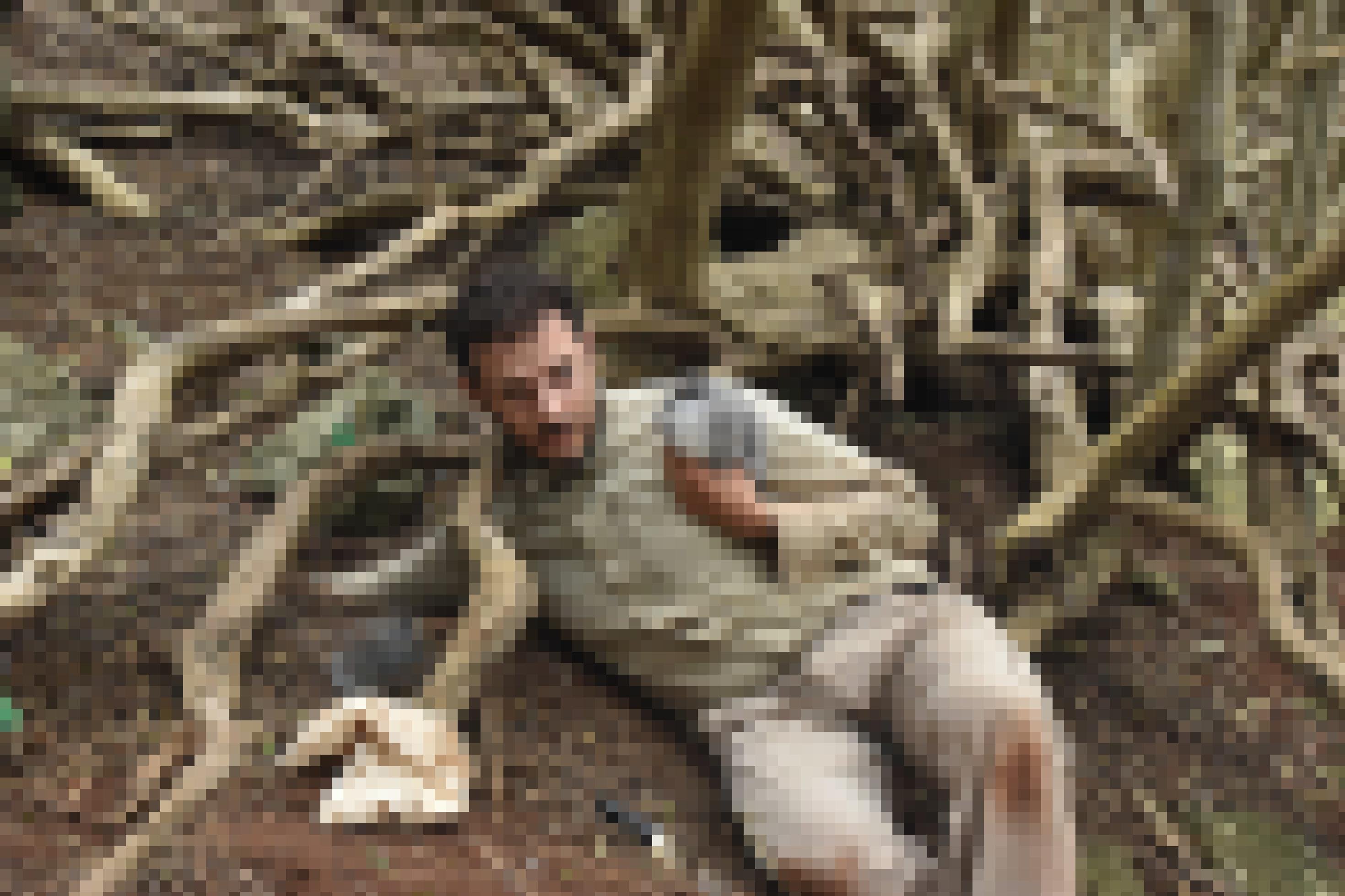 Foto des Forschers Luke Halpin in dreckiger Outdoor-Kleidung. Er liegt auf dem Boden in freier Natur, hält ein Vogelküken und greift mit dem anderen Arm in ein Loch.