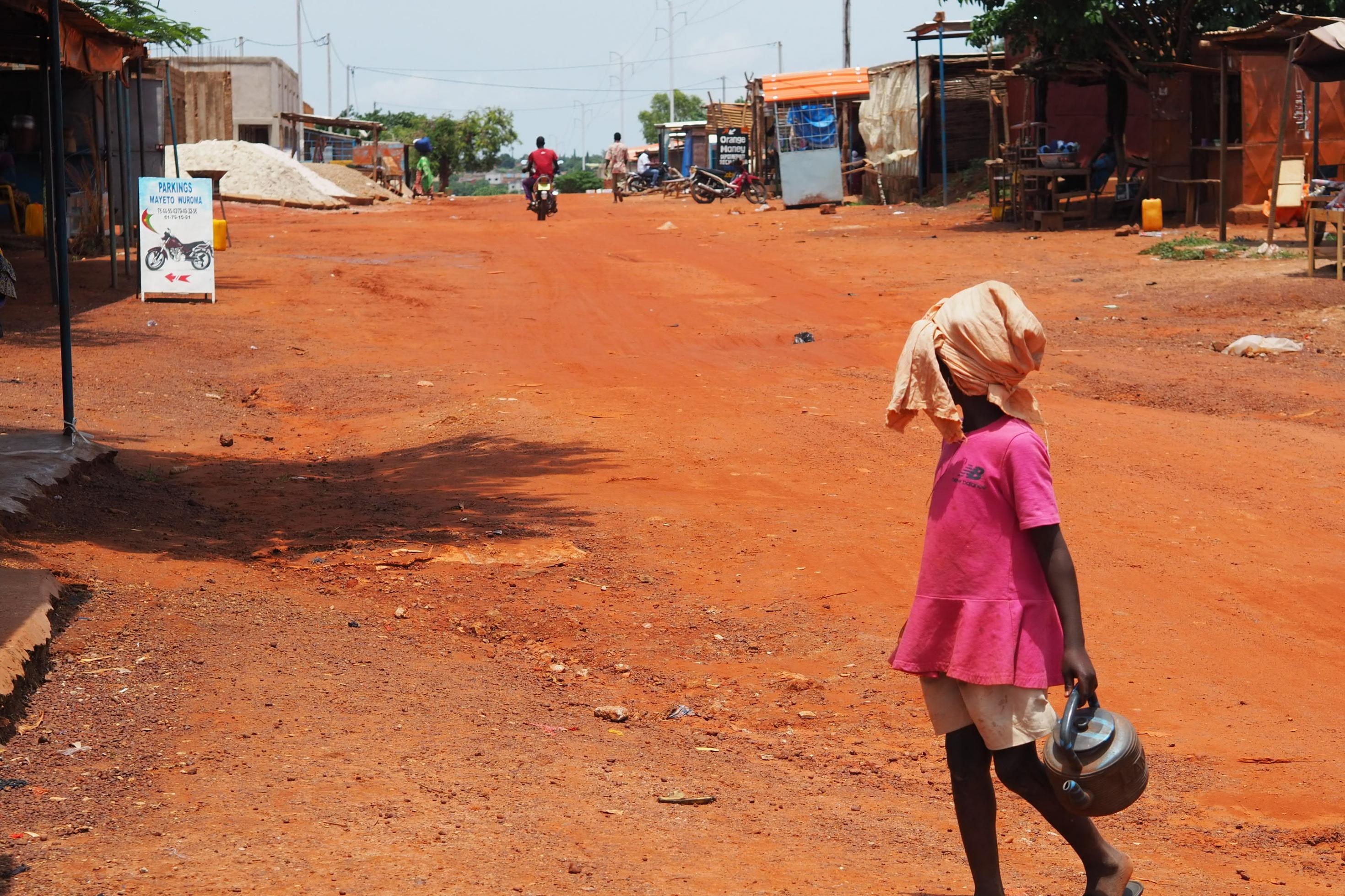 Ein Mädchen in Burkina Faso, Westafrika, geht mit einem Gebetswaschkessel auf einer staubigen Straße.