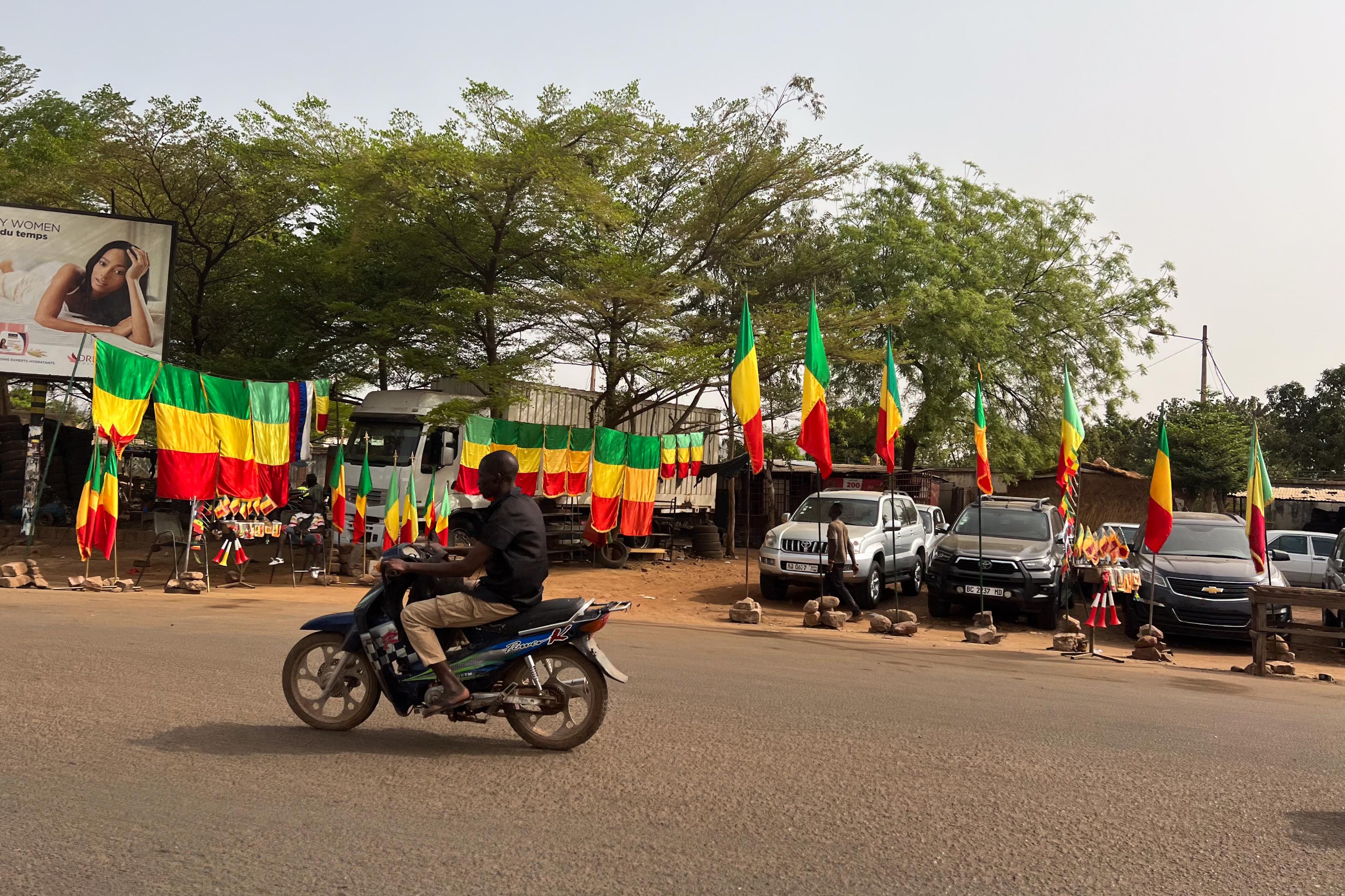 Am Rand einer größeren Straße sind etliche malische Flaggen zu sehen, sie werden zum Verkauf angeboten. Die Farben: grün-gelb-rot quer gesteift. Im Vordergrund fährt ein Mann auf einem Scooter vorbei.