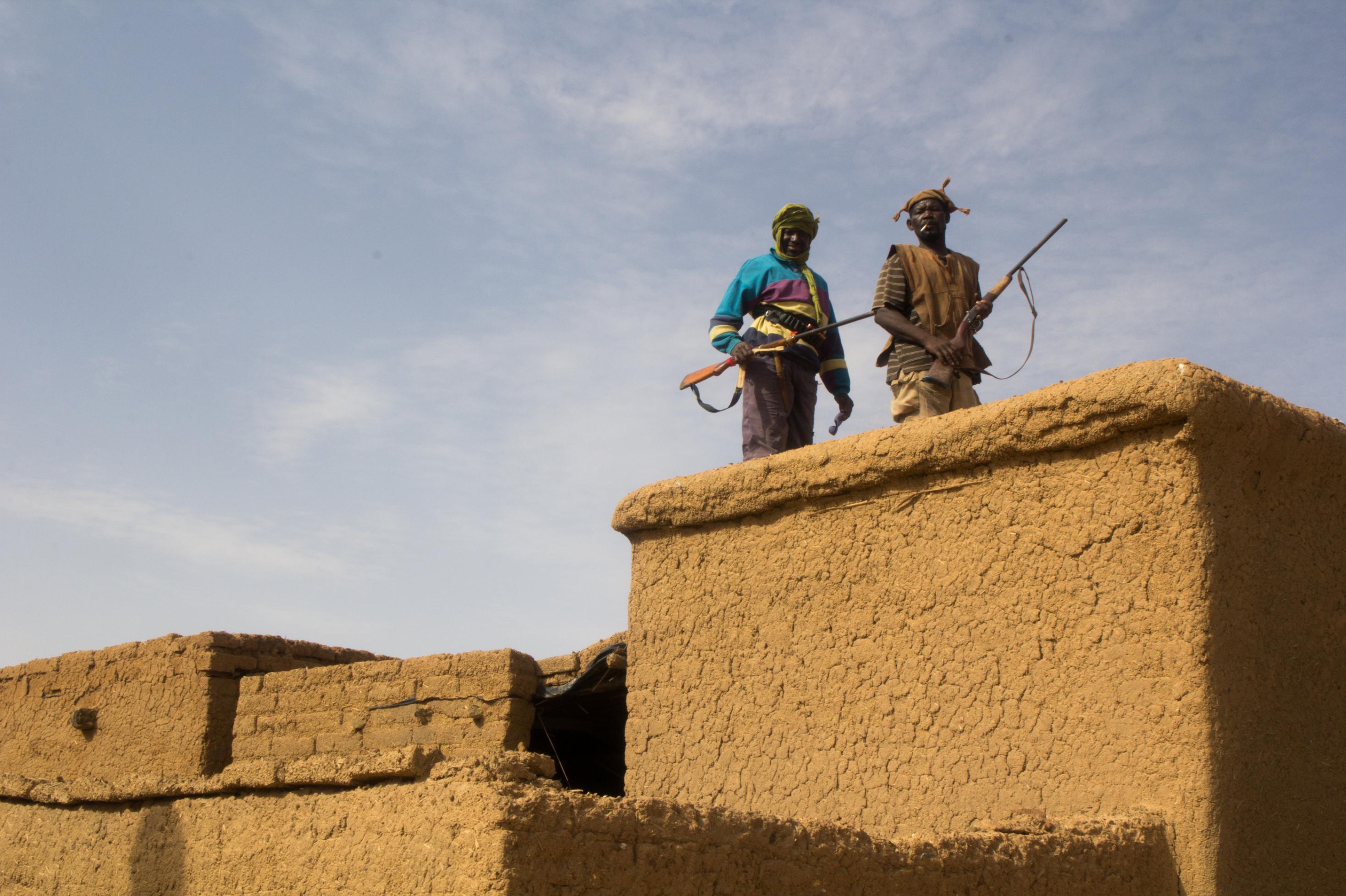 Auf dem Dach eines traditionellen Lehmhauses stehen zwei Männer mit Jagdgewehren. Sie tragen traditionelle Kleidung und Hüte aus grob gewebtem Stoff.